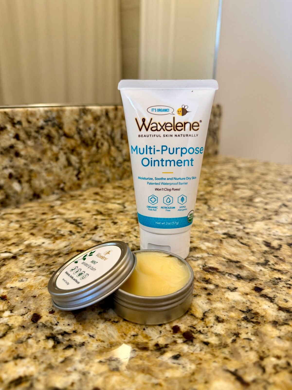 A tube of Waxelene Multi-Purpose Ointment stands upright on a granite countertop next to an open metal tin containing a balm, with a bathroom mirror in the background.