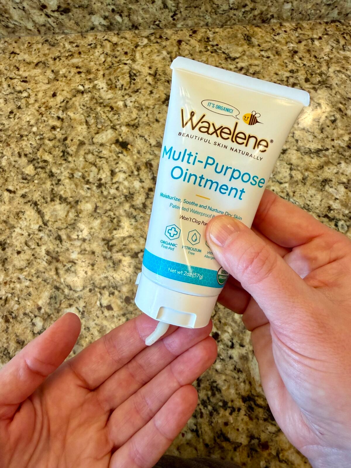 A person dispenses Waxelene Multi-Purpose Ointment from a white tube onto their hand over a granite countertop.