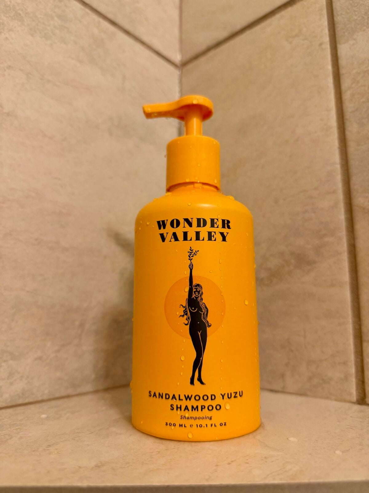 A yellow pump bottle of Wonder Valley Sandalwood Yuzu Shampoo sits on the corner of a tiled shower shelf, with water droplets visible on the bottle.
