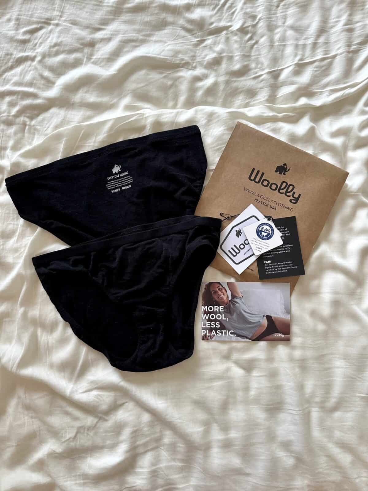 Black Woolly merino underwear laid out on a bed with packaging, product tags, and a postcard that reads 'More wool, less plastic.