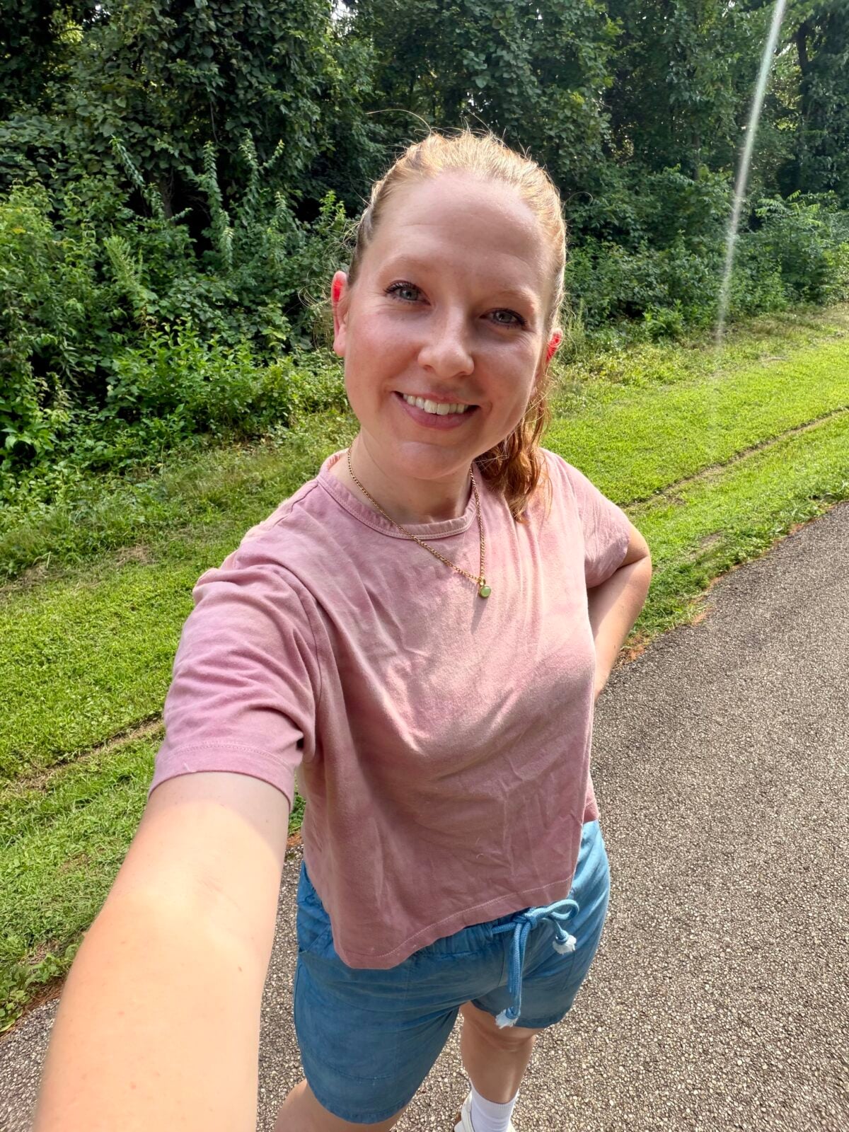 A woman stands on a paved path next to grass and trees, taking a selfie. She is wearing a pink T-shirt, blue shorts, and a necklace, and is smiling at the camera on a sunny day.