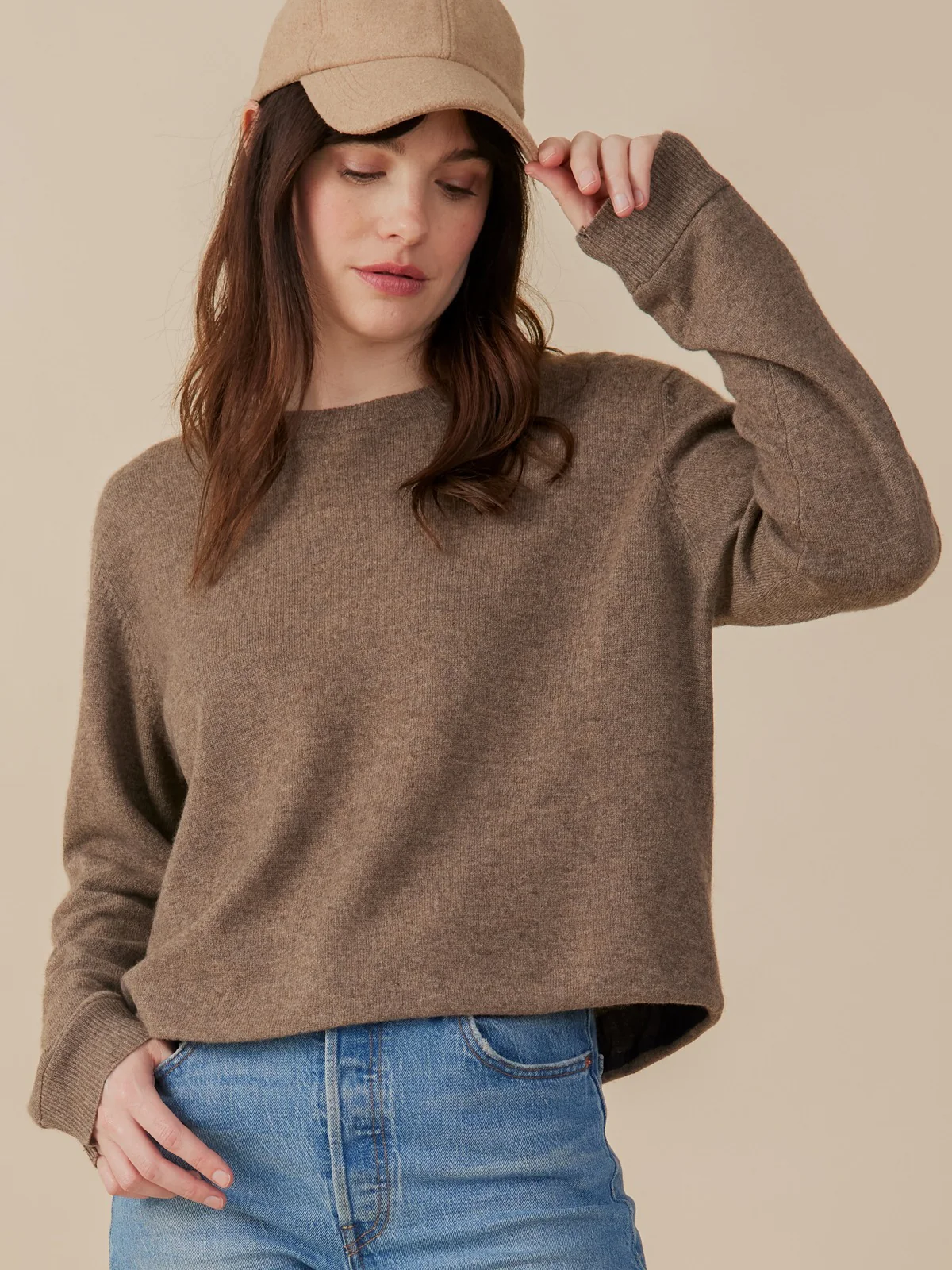 A model wearing a relaxed-fit crewneck sweater in heathered brown, styled with blue jeans and a tan baseball cap.