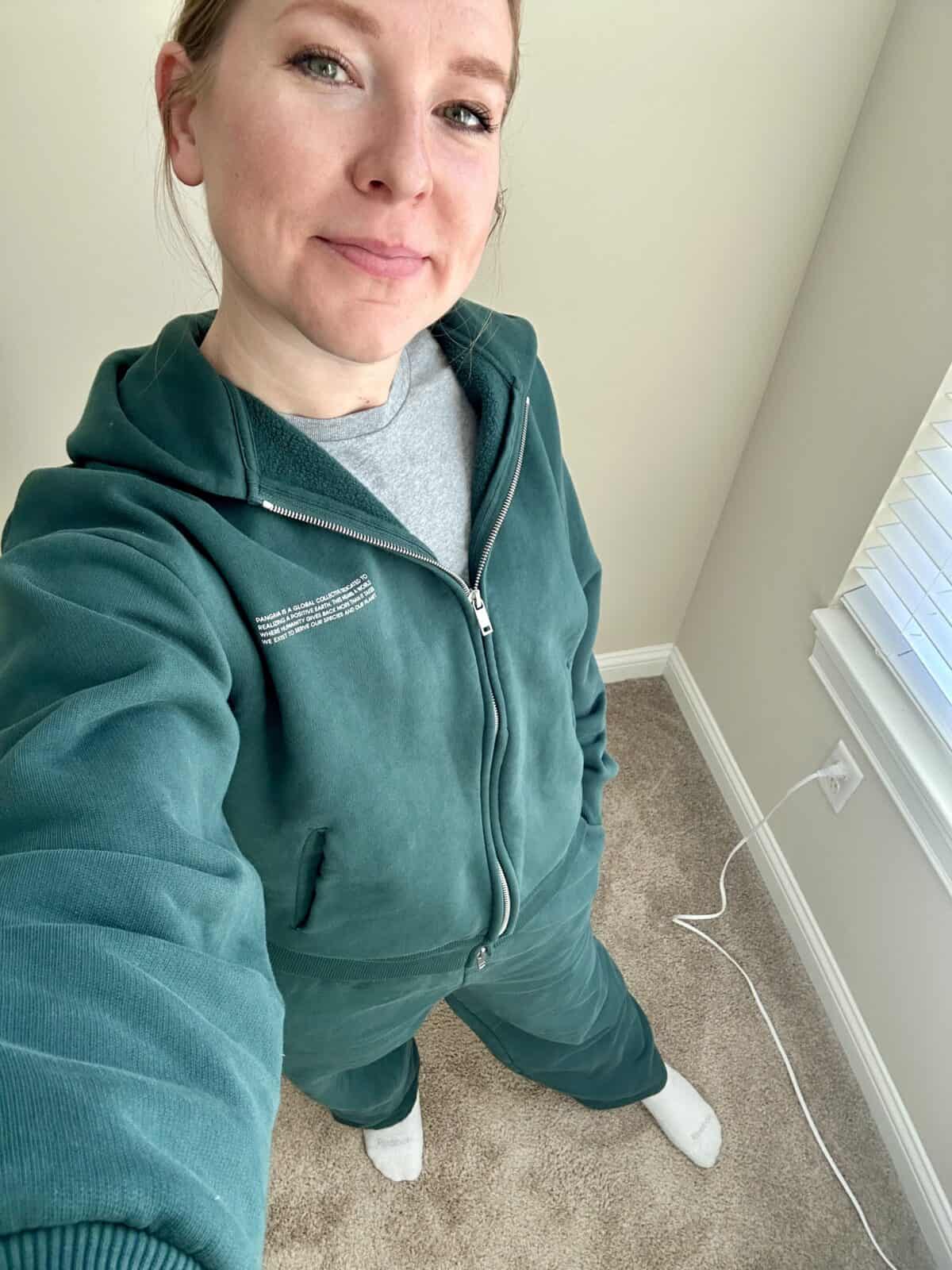 A person standing indoors wearing a matching green zip-up hoodie and sweatpants with a gray T-shirt and white socks. The photo is taken from a slightly elevated angle, showing beige carpet, a wall outlet, and a window with blinds.