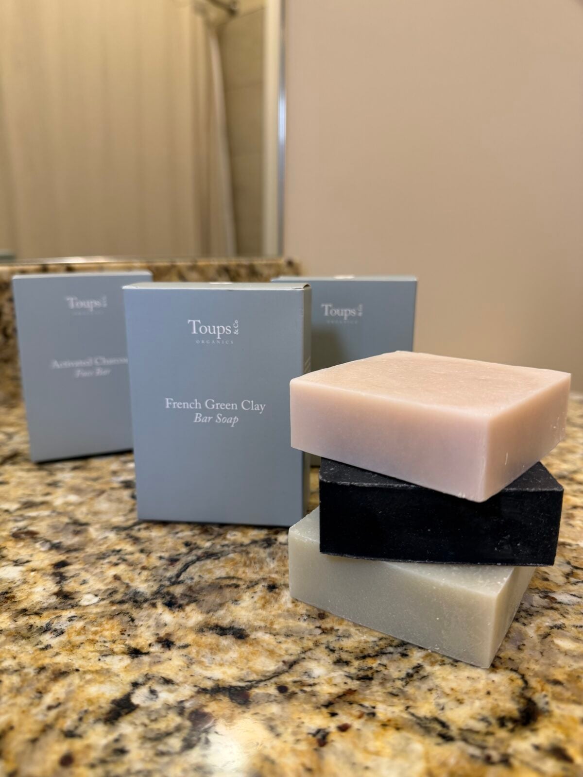 Three bar soaps—one light pink, one black, and one light green—are stacked on a granite countertop. Behind them are two Toups & Co. soap boxes labeled French Green Clay and Activated Charcoal.