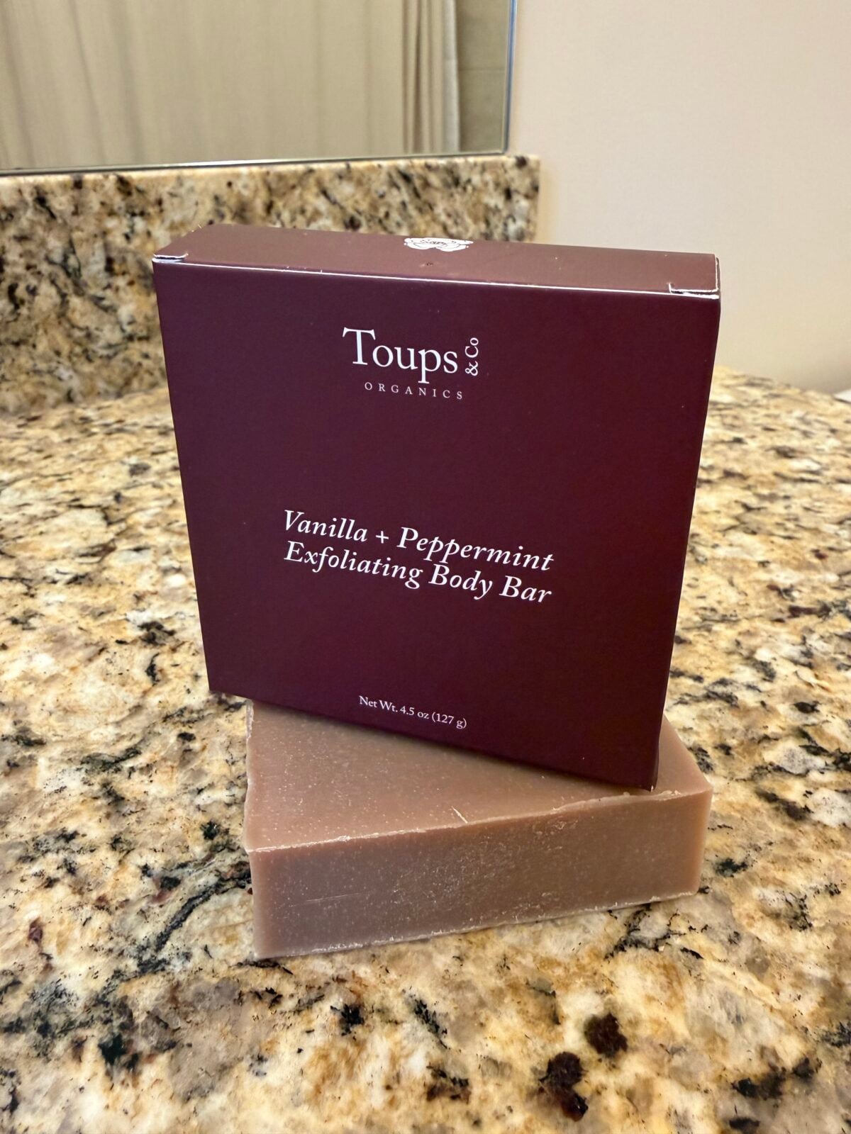 A box labeled &ldquo;Toups & Co Organics Vanilla + Peppermint Exfoliating Body Bar&rdquo; sits on a granite countertop, with a bar of soap placed beneath it. A mirror and part of a sink are in the background.