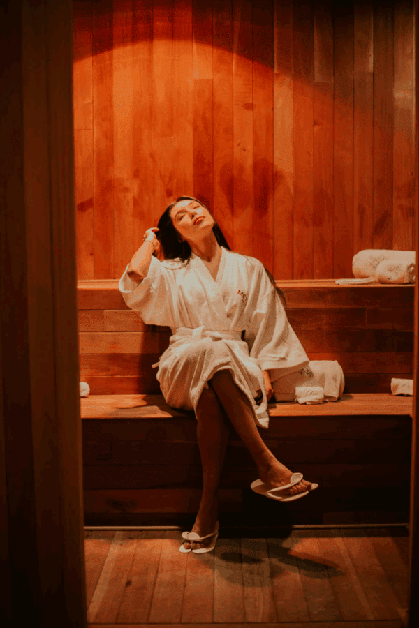 Woman in a white robe sitting peacefully in a warmly lit sauna, leaning back and relaxing.