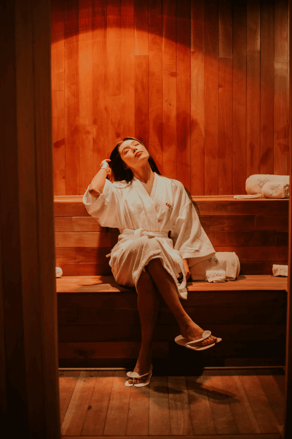 Woman in a white robe sitting peacefully in a warmly lit sauna, leaning back and relaxing.