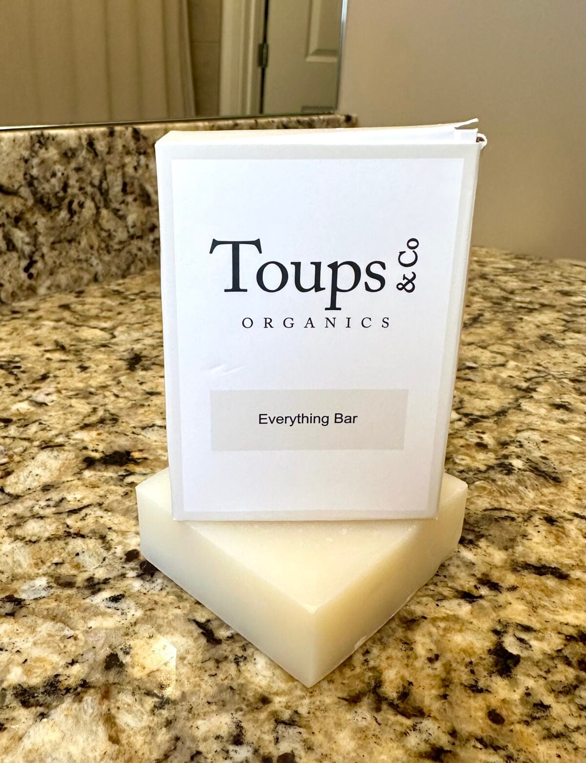 Toups & Co Organics Everything Bar soap displayed on a granite bathroom counter with its white packaging behind it.