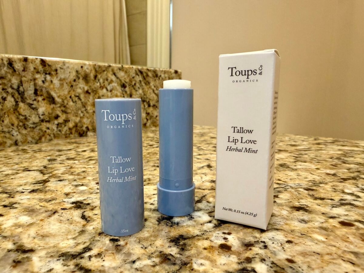 A blue tube of Toups Organics Tallow Lip Love Herbal Mint lip balm stands next to its packaging on a granite bathroom counter with a mirror in the background.