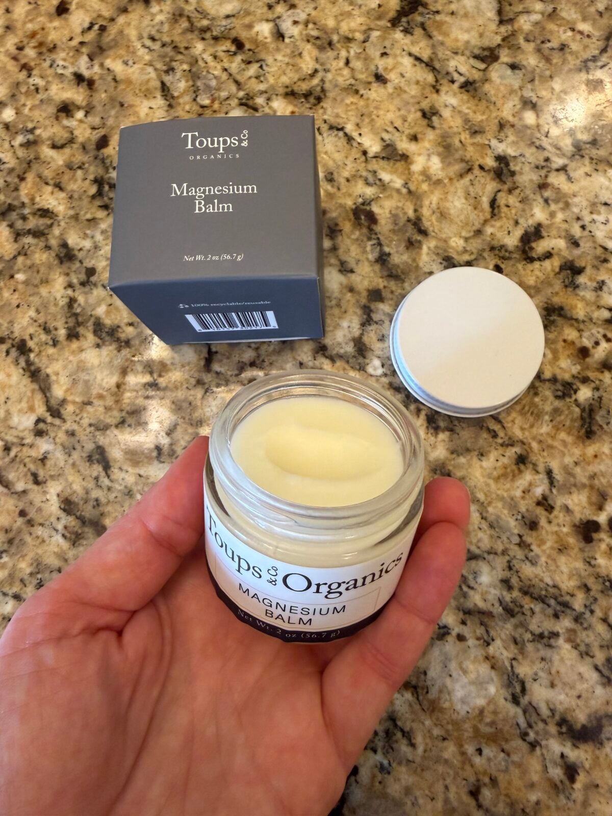 A hand holds an open jar of Toups & Co. Organics Magnesium Balm above a granite countertop. The jars lid and the products box sit nearby on the counter.