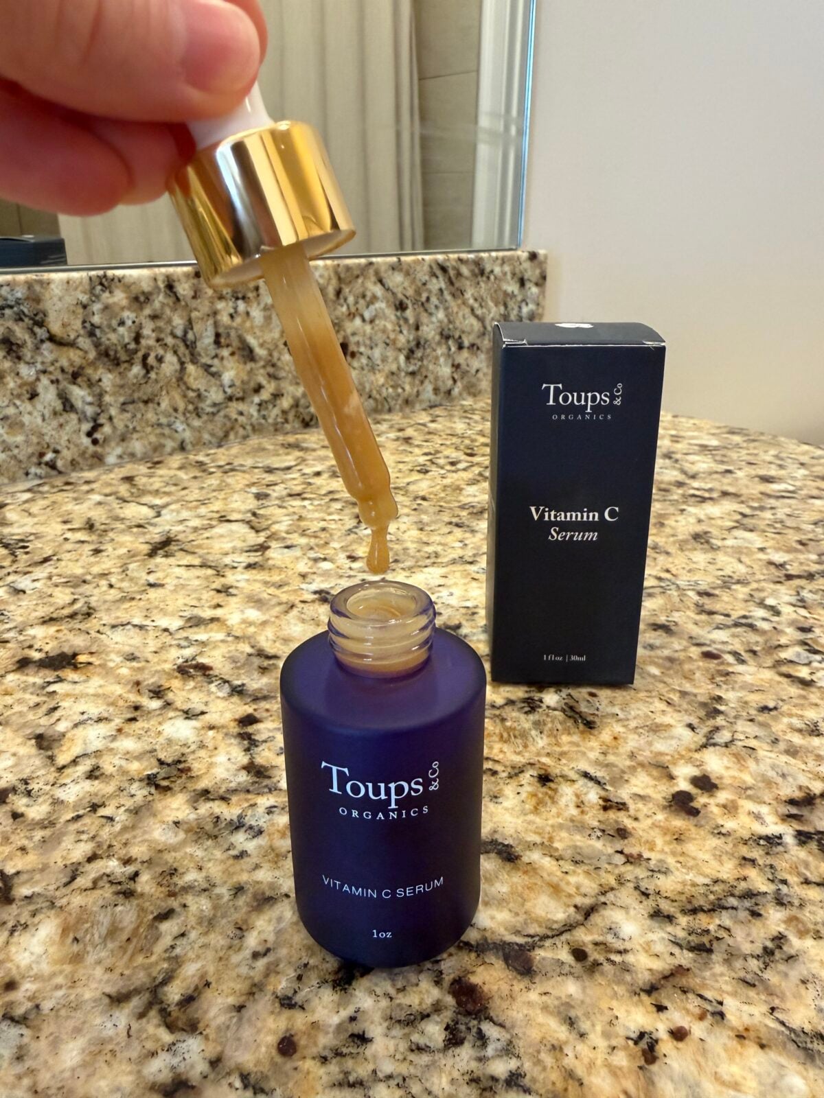 Toups & Co Organics Vitamin C Serum bottle with dropper dispensing golden serum, navy box in background on granite counter