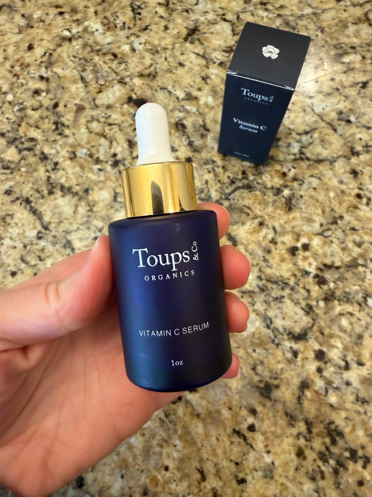 Hand holding Toups & Co Organics Vitamin C Serum bottle with gold dropper cap and white rubber bulb, navy box visible behind