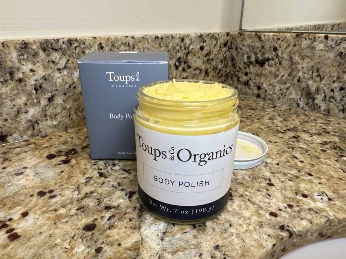 Open jar of Toups & Co Organics Body Polish with yellow scrub inside, sitting on a granite bathroom counter next to its gray product box.