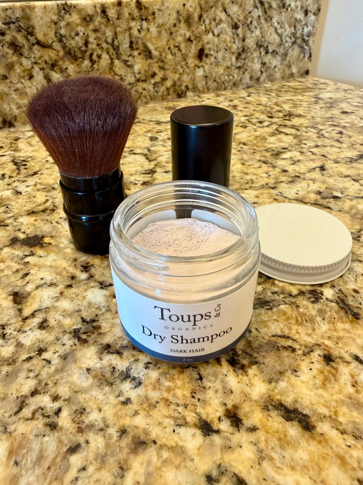 A jar of Toups & Co Organics Dry Shampoo for dark hair sits open on a granite countertop, with its lid off to the side. A makeup brush and a black cylindrical container are positioned nearby.