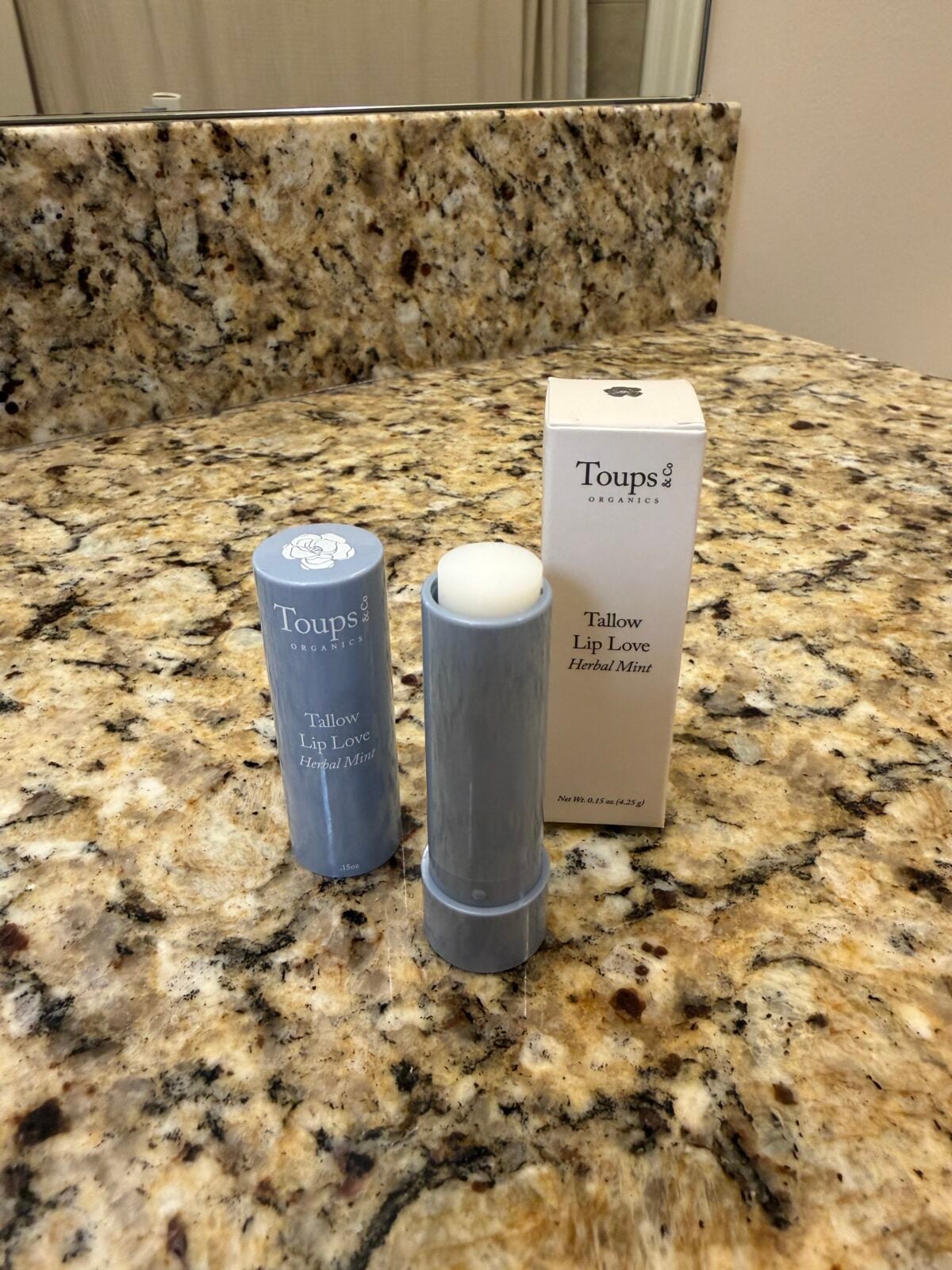 A tube of Toups & Co. Tallow Lip Love Herbal Mint lip balm stands open on a granite bathroom countertop next to its cap and packaging box. The background shows a bathroom sink area.
