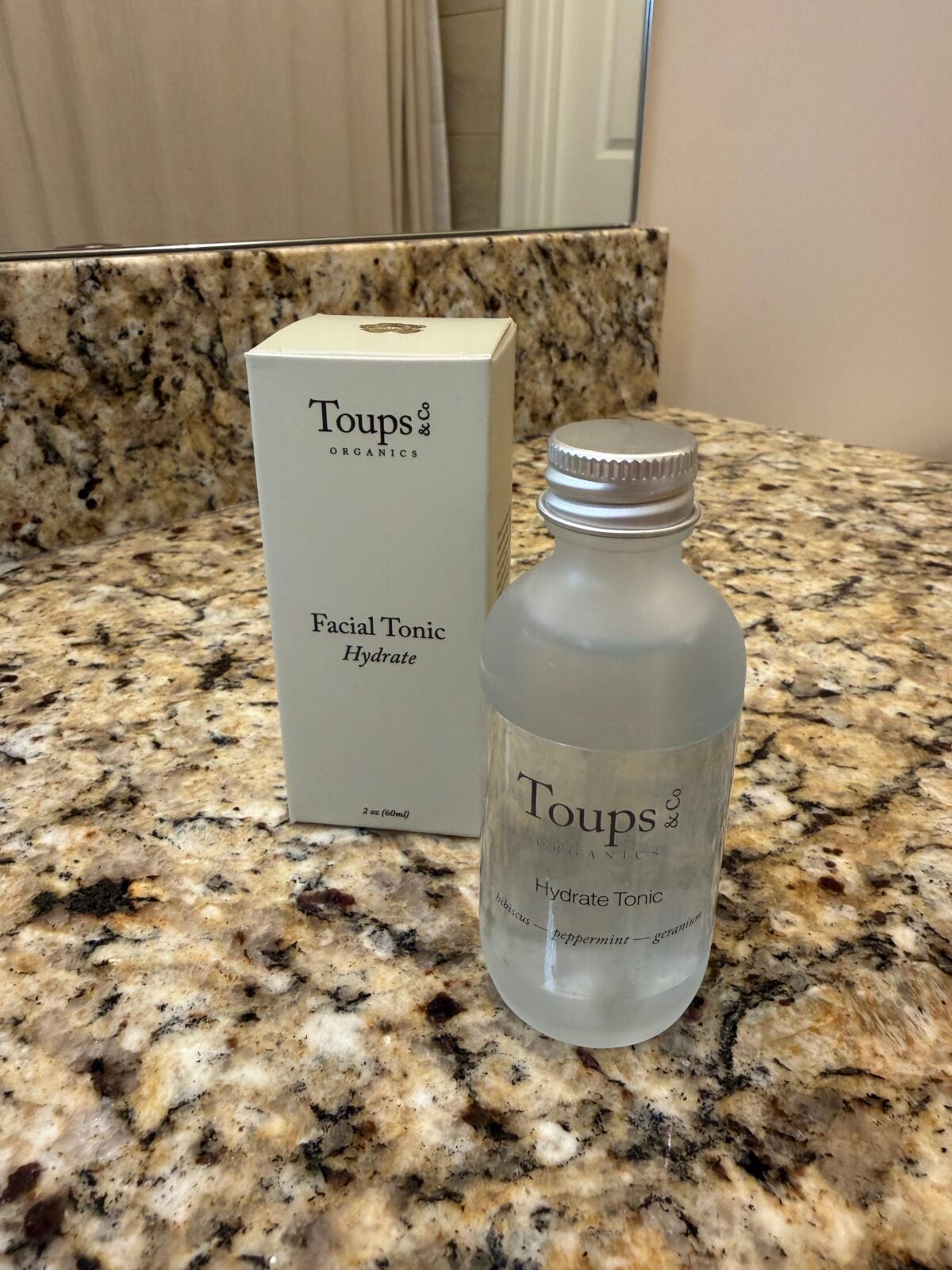Toups & Co Organics Hydrate Facial Tonic bottle with aluminum cap and box, 2 oz, with hibiscus, peppermint, and geranium, on granite bathroom counter