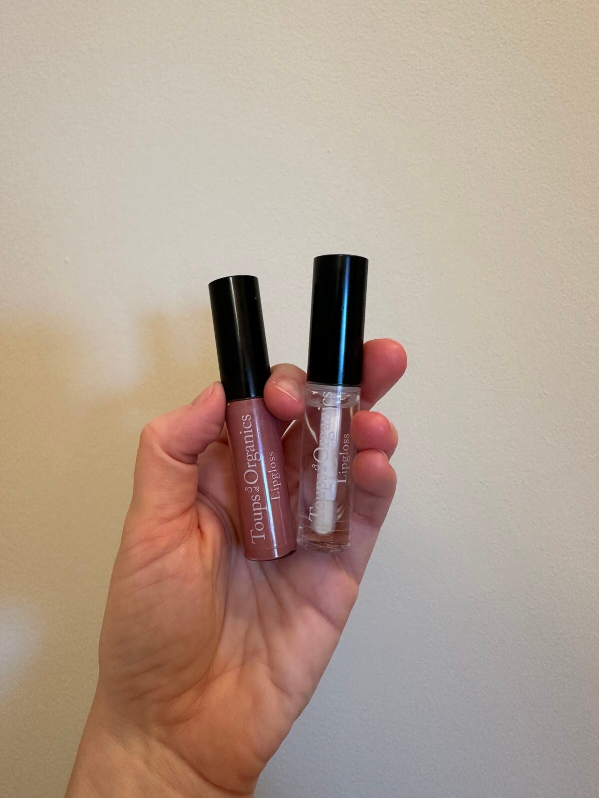 A hand holds two tubes of Tropic Organics lip products, one with a pink shade and the other clear, both with black caps, against a plain beige background.