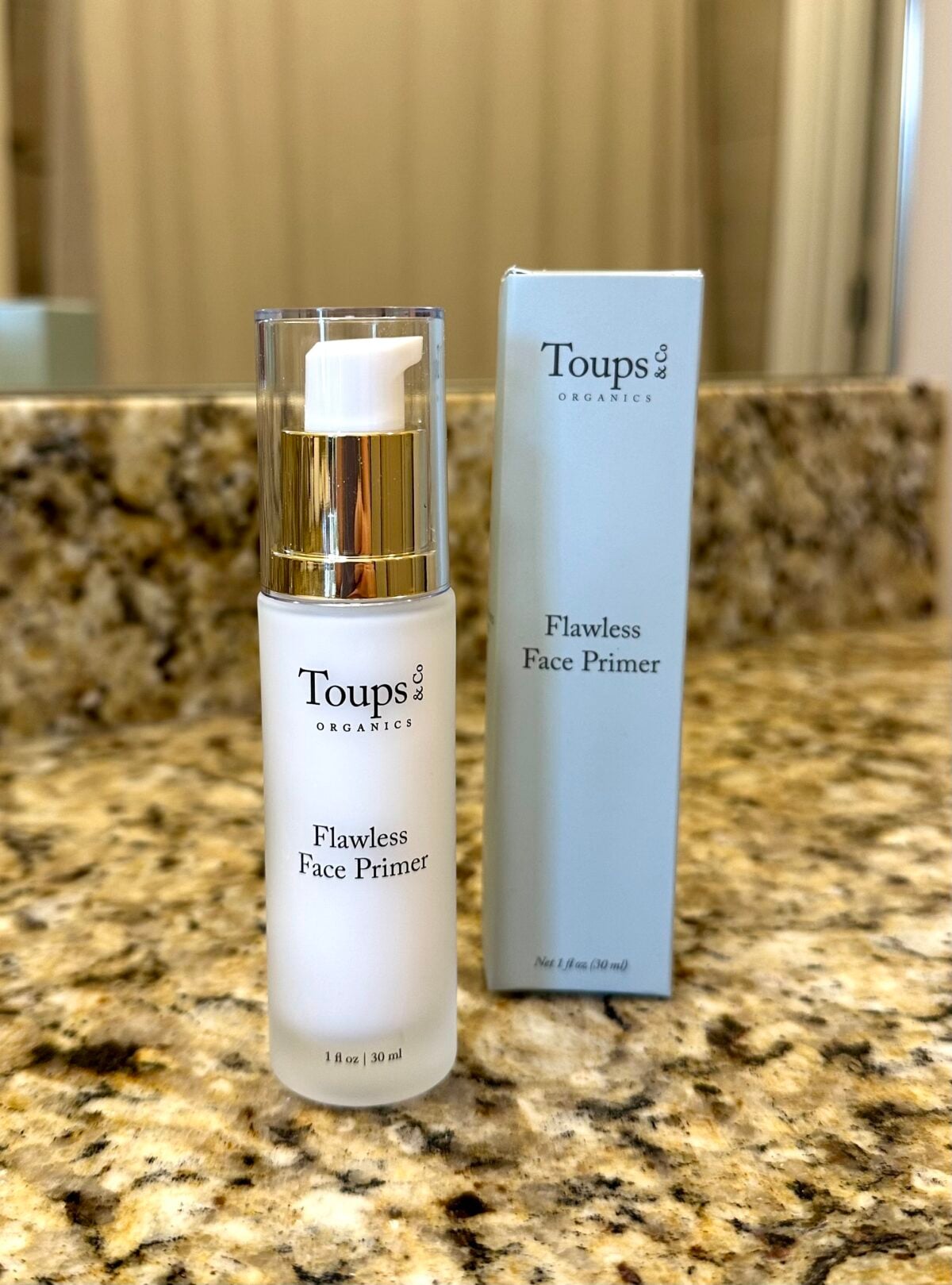 Bottle of Toups & Co Flawless Face Primer standing next to its box on a granite countertop.
