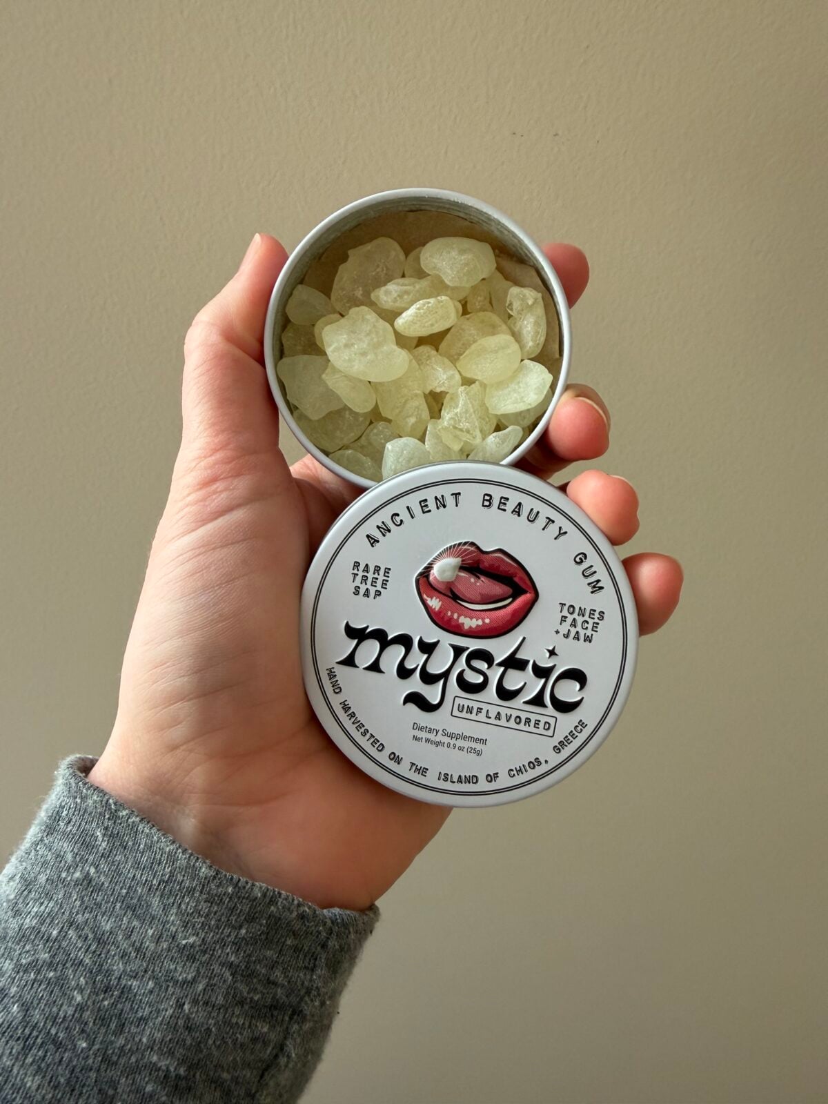 Hand holding open tin of Mystic unflavored mastic gum showing translucent pale yellow gum pieces inside