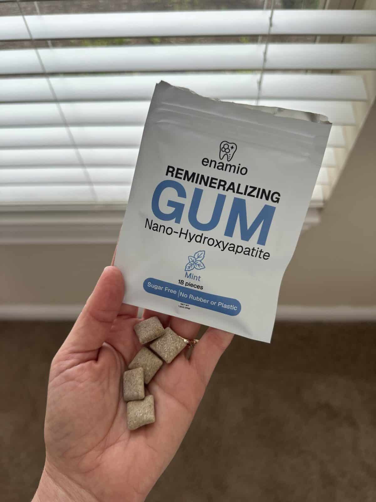 A hand holding several square pieces of beige chewing gum in front of a white package labeled &ldquo;enamio Remineralizing Gum &ndash; Nano-Hydroxyapatite, Mint, 18 pieces, Sugar Free, No Rubber or Plastic.&rdquo; The background shows a window with blinds.