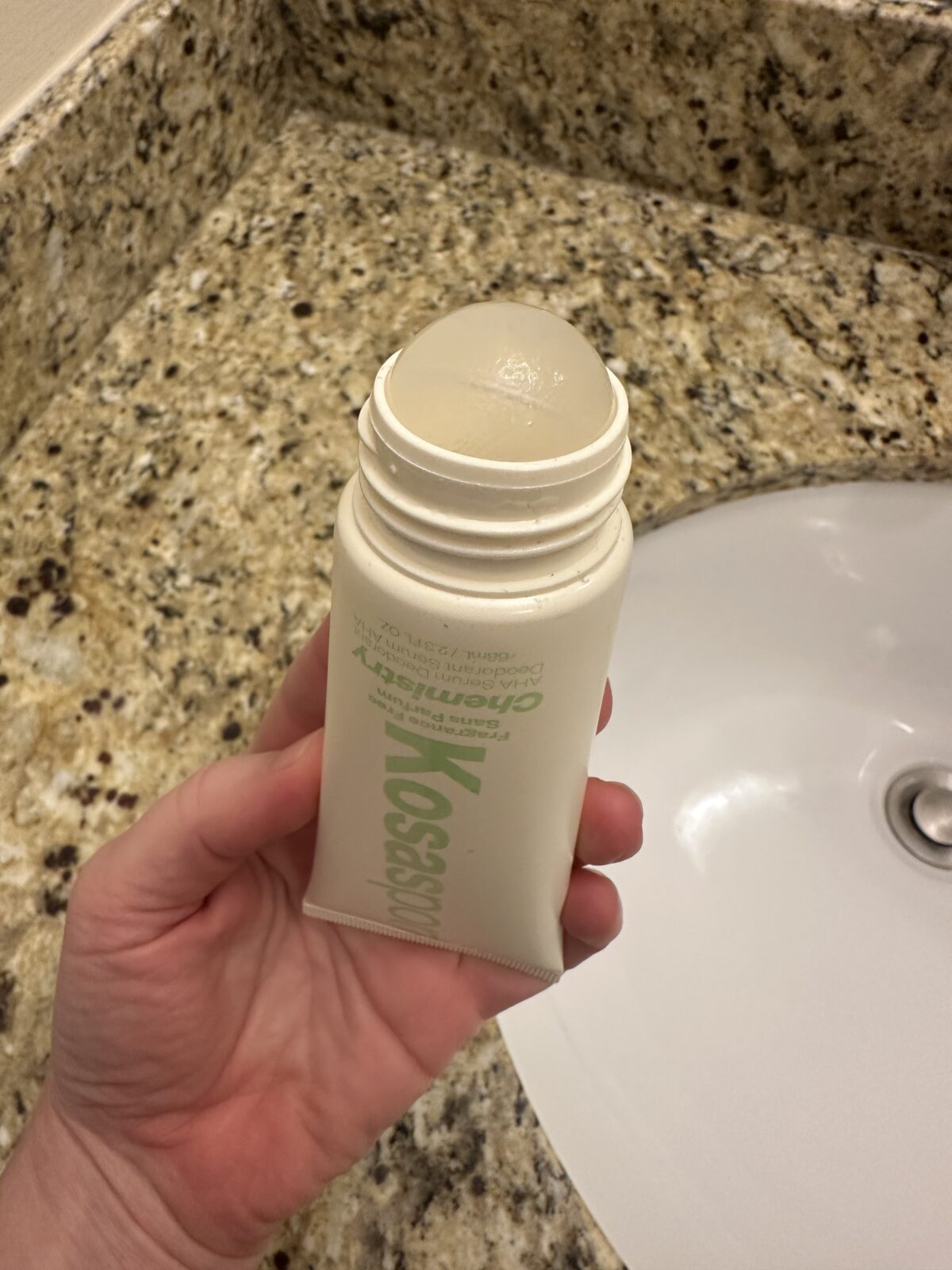 Hand holding an open Kosas Chemistry deodorant tube with the rounded gel deodorant exposed over a granite bathroom counter and sink.