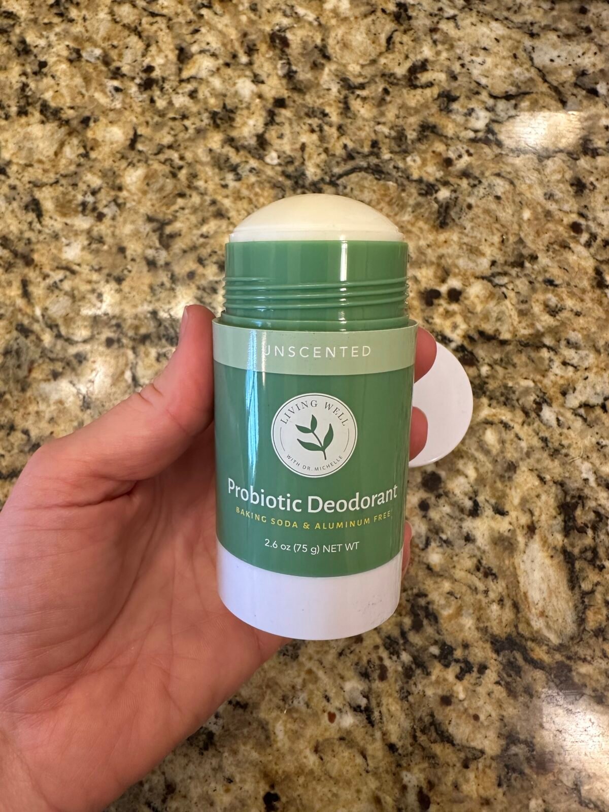 Hand holding an open Living Well with Dr. Michelle Unscented Probiotic Deodorant stick over a speckled granite countertop.