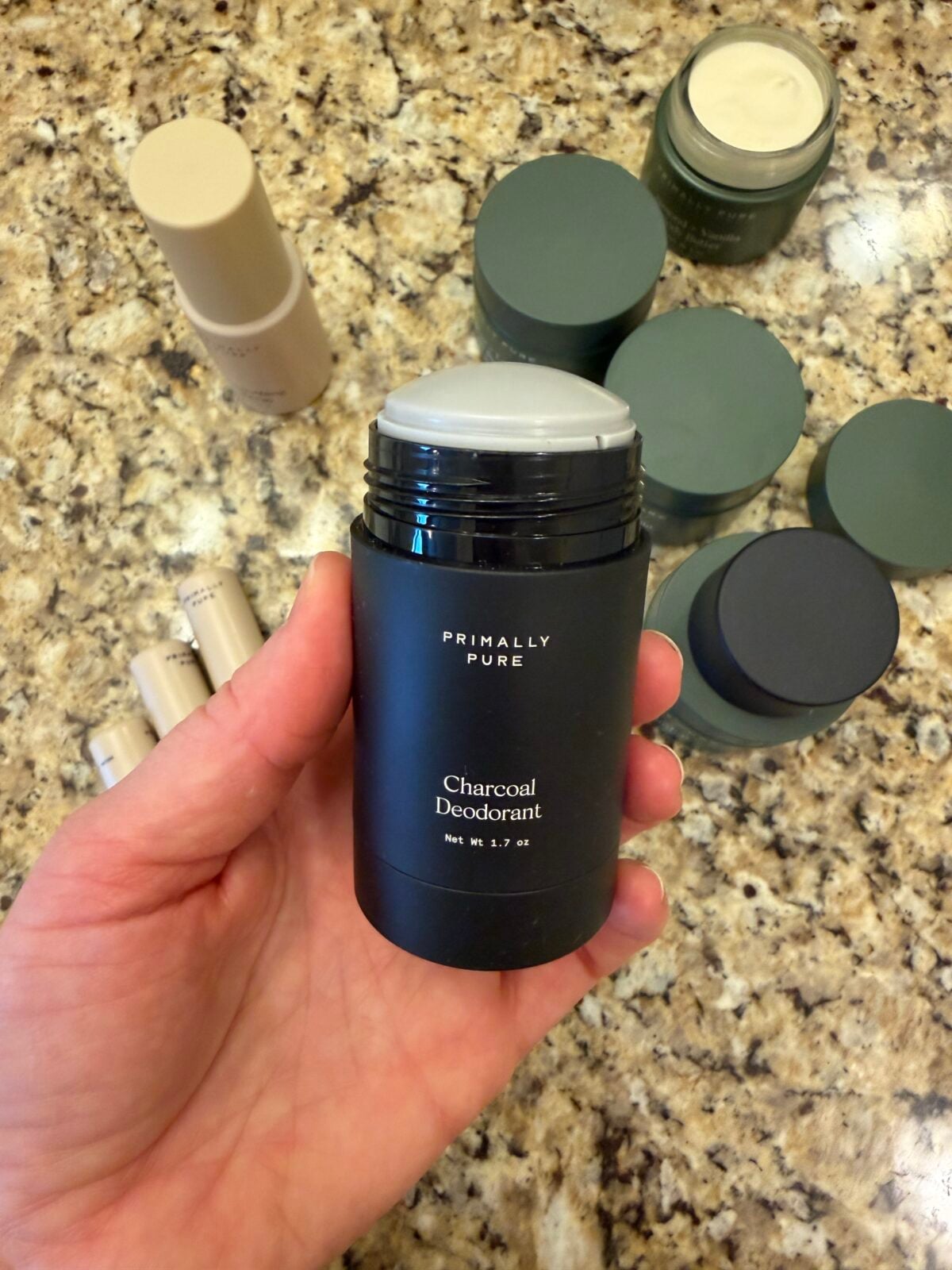 Hand holding Primally Pure Charcoal Deodorant stick with multiple natural deodorant products in background