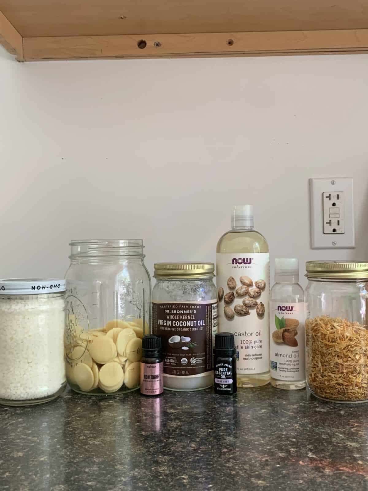 A countertop is shown with an array of jars and bottles containing ingredients such as castor oil, almond oil, coconut oil, cocoa butter wafers, and what appears to be calendula.