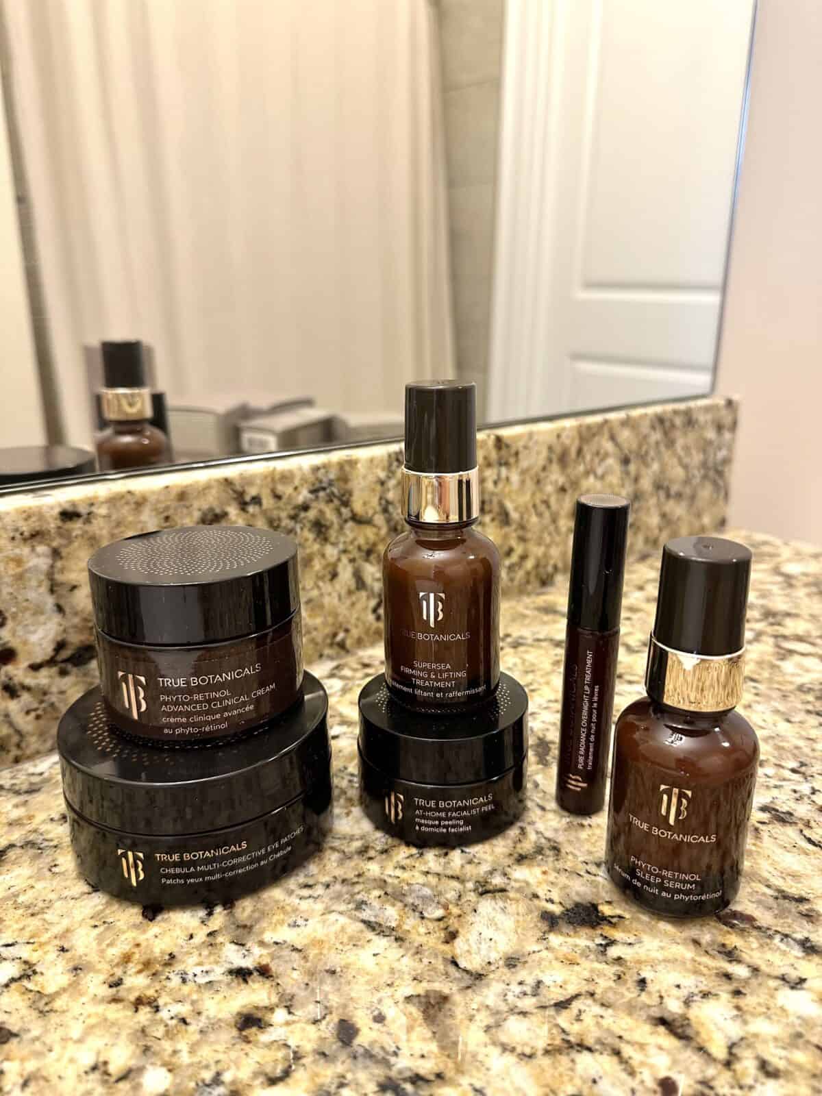 Collection of True Botanicals skincare products in brown bottles and jars displayed on a bathroom counter.