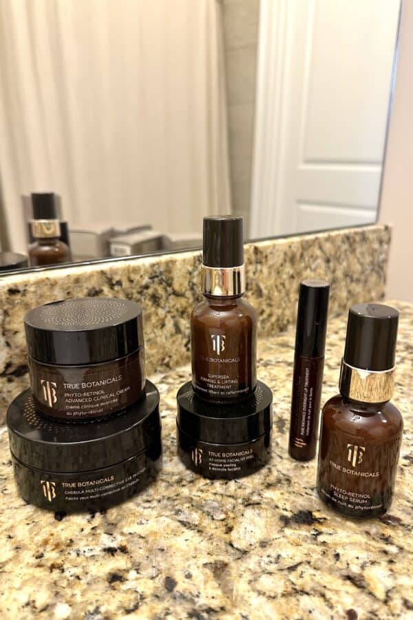 Collection of True Botanicals skincare products in brown bottles and jars displayed on a bathroom counter.