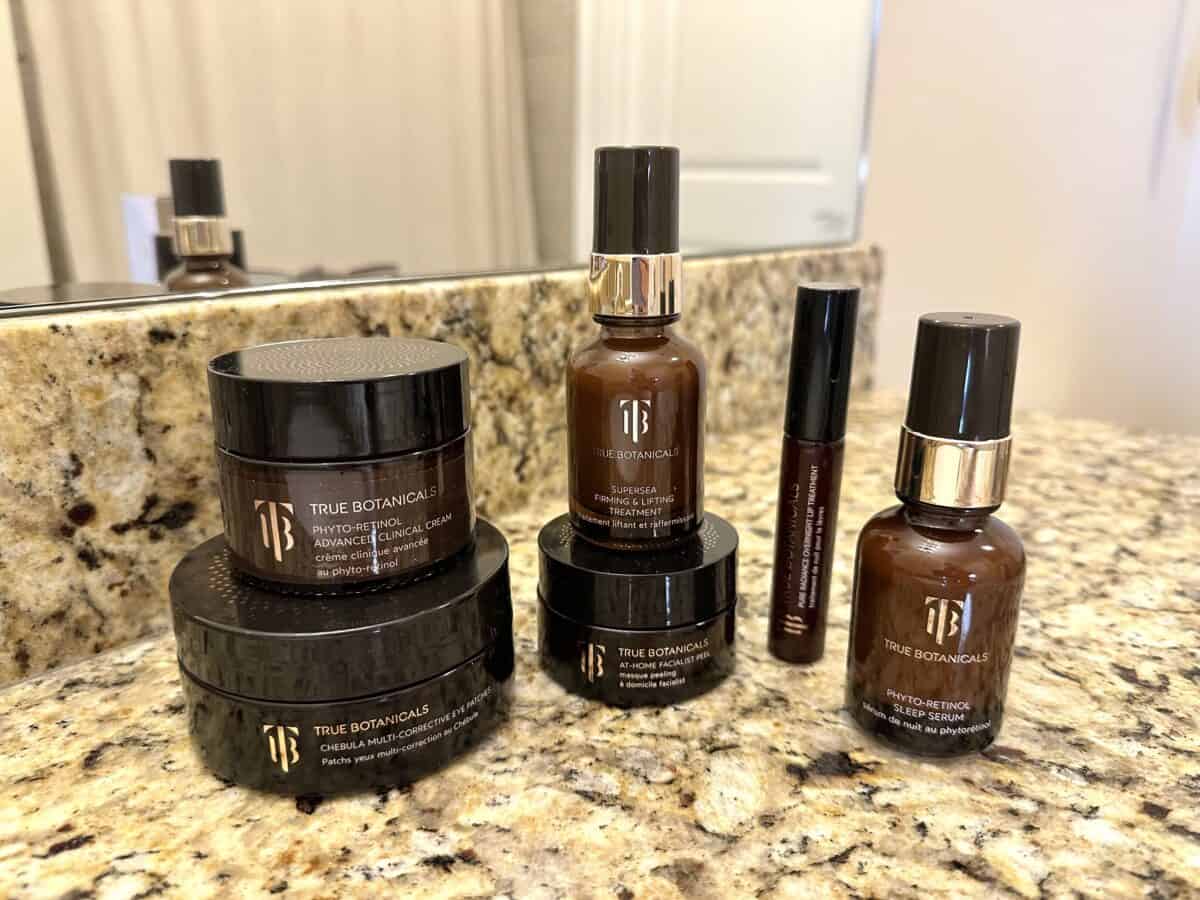 Collection of True Botanicals skincare products in brown bottles and jars displayed on a bathroom counter.