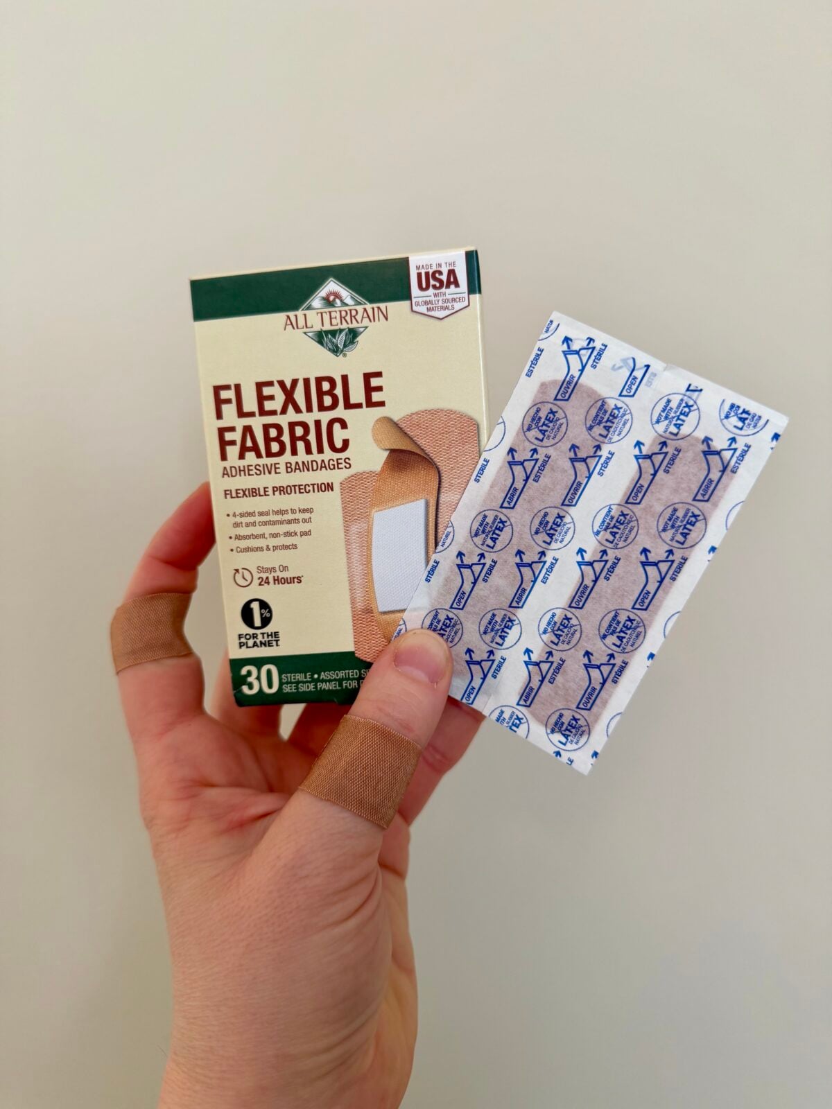 A hand with two bandaged fingers holds a box of All Terrain Flexible Fabric adhesive bandages and a single wrapped bandage against a plain background.