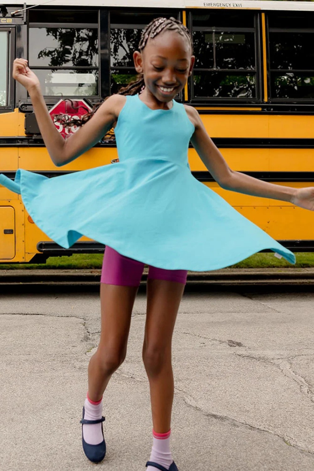 A smiling girl wearing a light blue dress and purple shorts spins in front of a yellow school bus on a paved street. She has braided hair and is wearing socks and blue shoes.