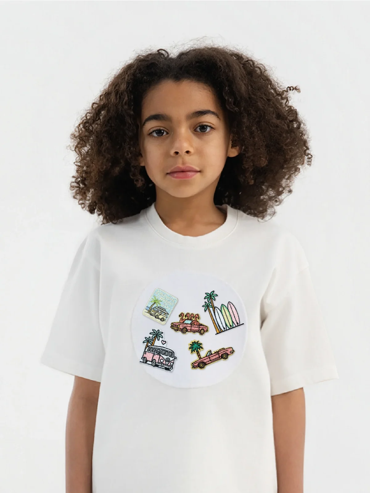 A child with curly hair wears a white T-shirt featuring colorful illustrations of cars, palm trees, buildings, and a postcard on the front. The background is plain and white.