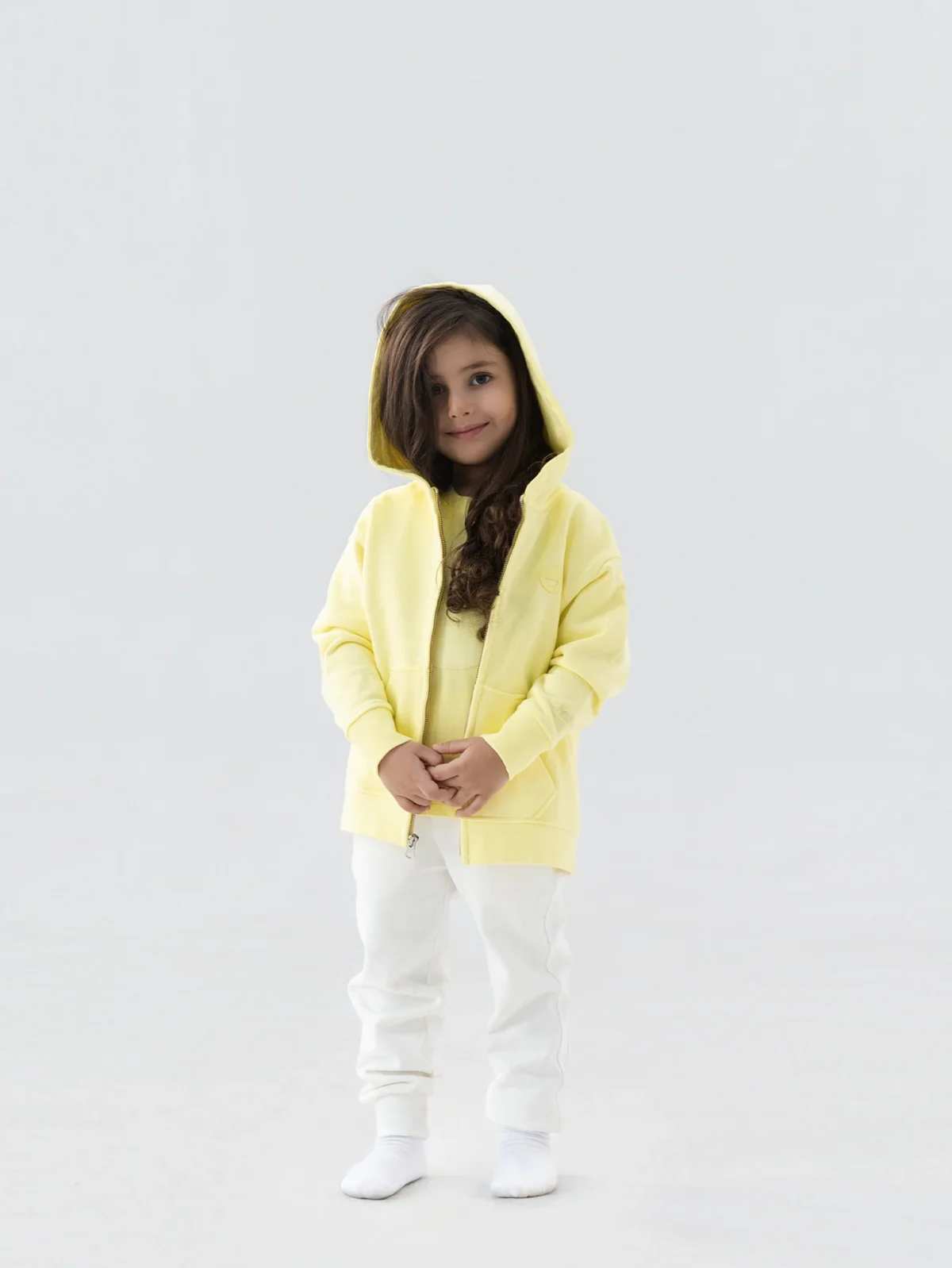A young child with long brown hair stands facing forward, wearing a yellow hoodie, white pants, and white socks, against a plain light grey background.