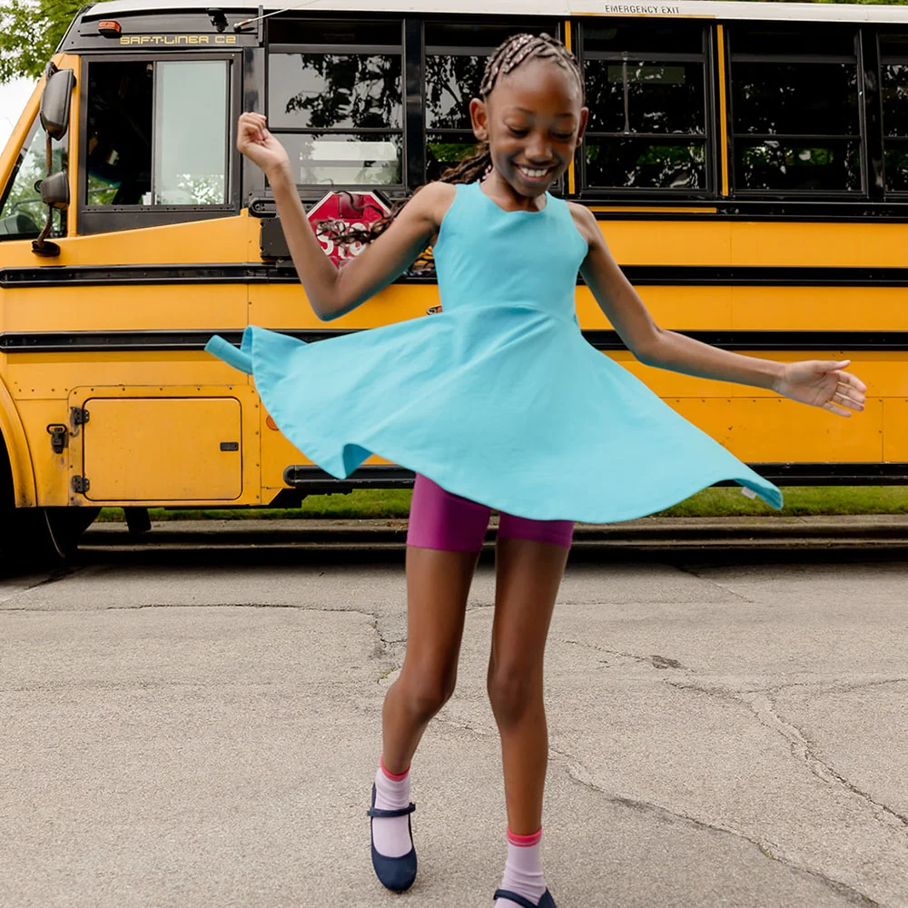 A child wearing a blue dress and purple shorts spins joyfully on a street, with a yellow school bus and trees in the background.