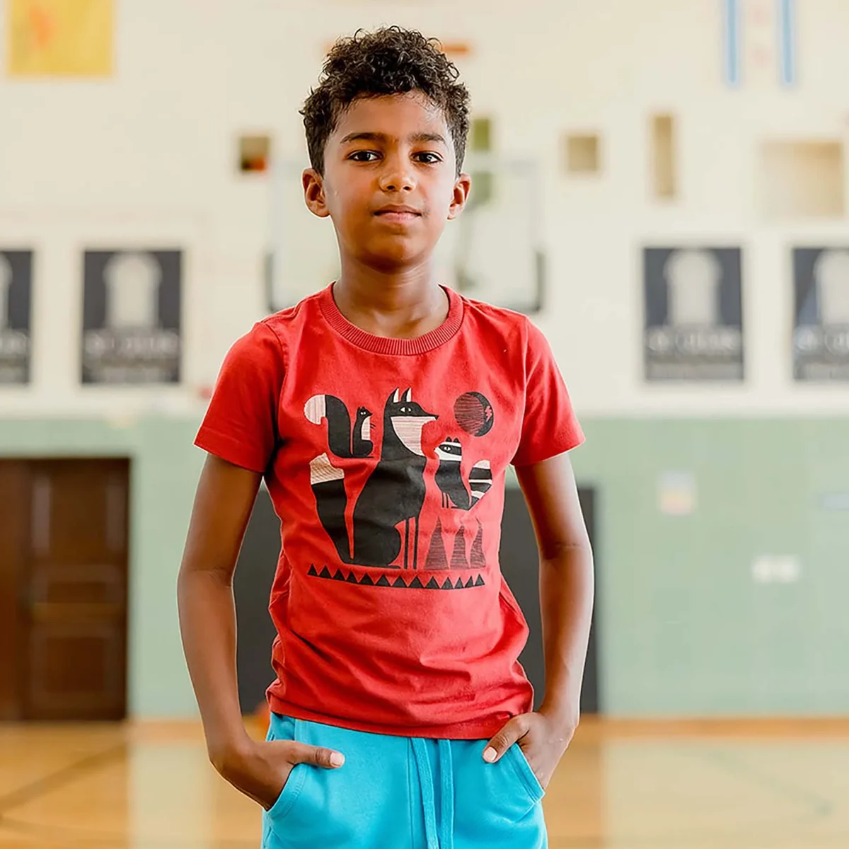 A boy stands indoors wearing a red t-shirt with a black and orange animal graphic and blue shorts. The background includes a gymnasium wall, double doors, and banners.