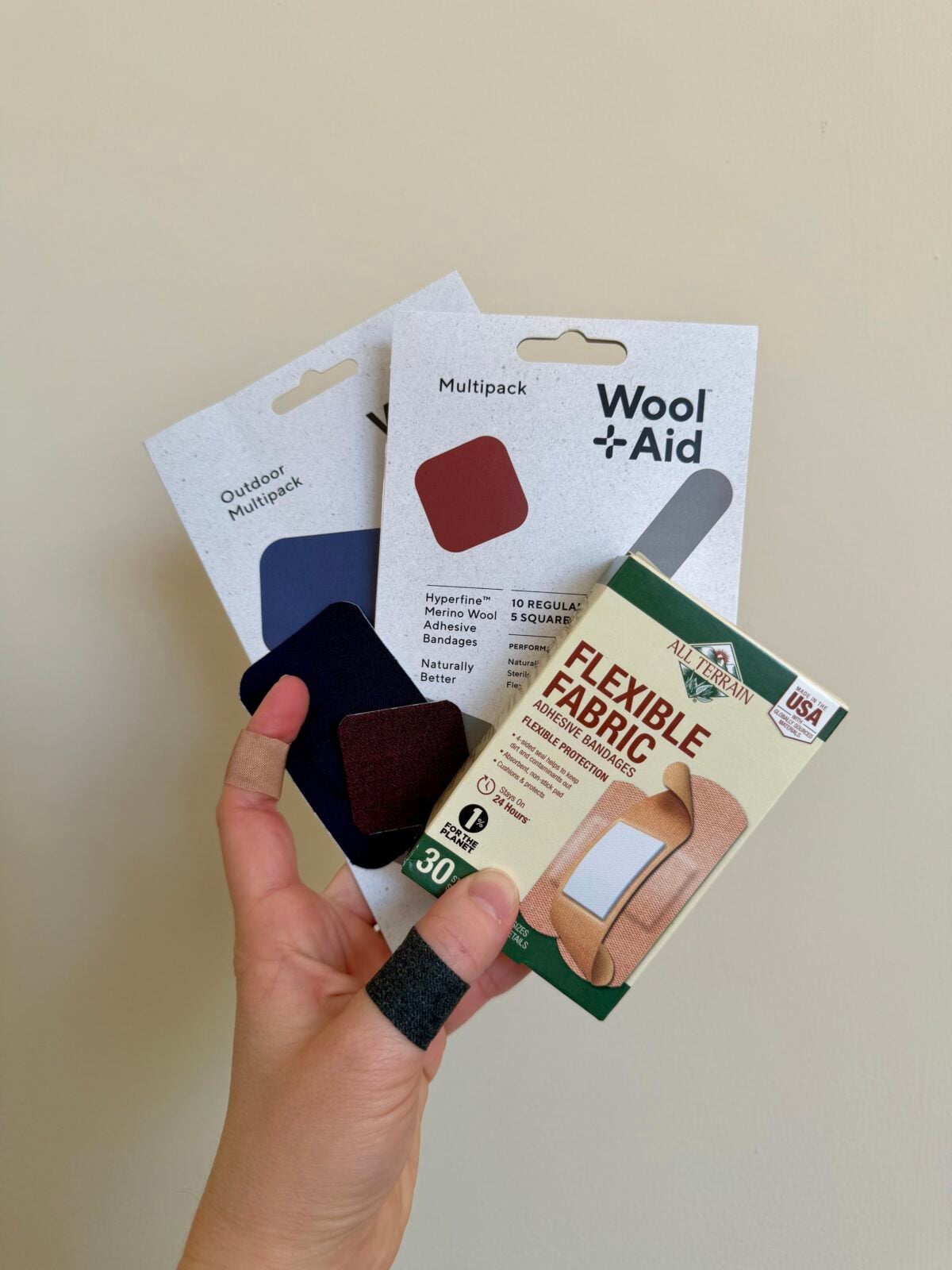 A hand with two fingers wrapped in black bandages holds two Wool Aid multipacks and a box of All Health flexible fabric bandages against a plain beige background.