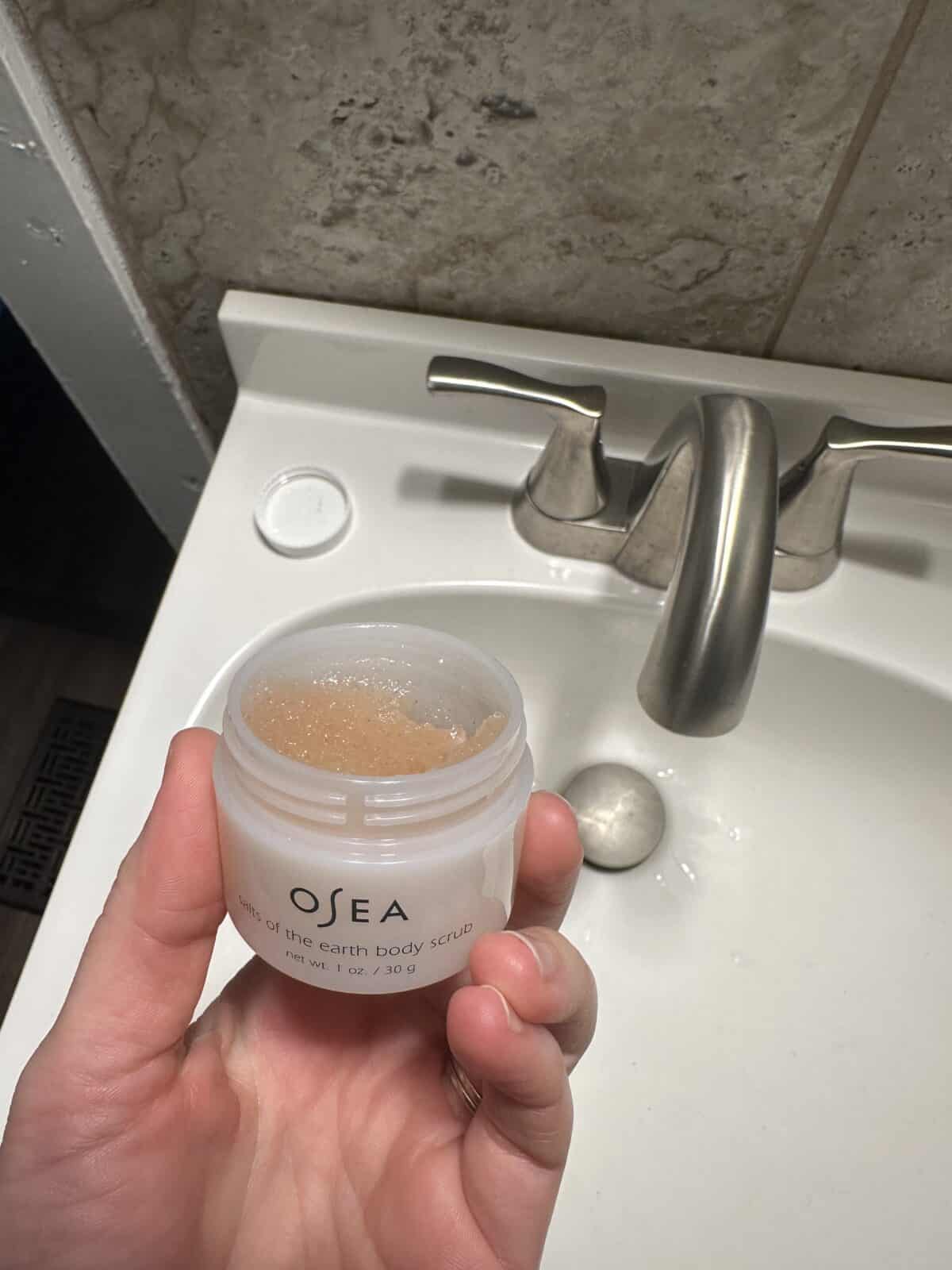 A person holding the OSEA Salts of the Earth Body Scrub close to the bathroom sink.