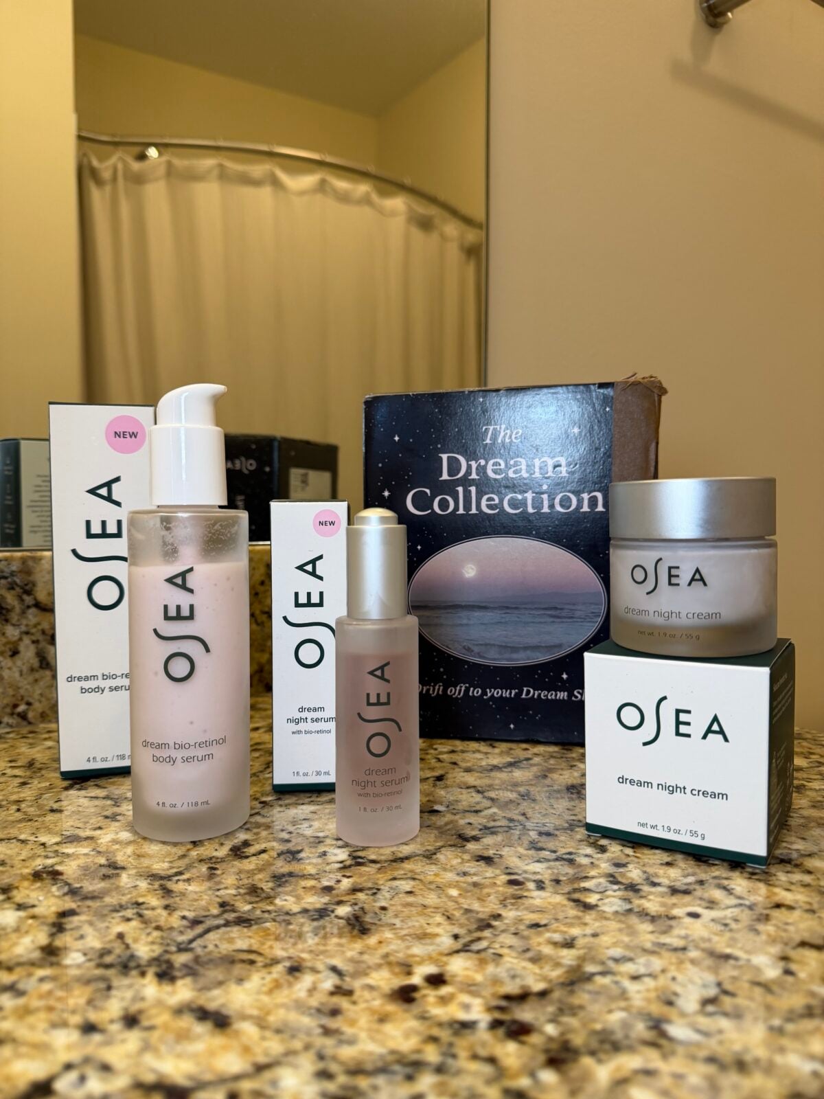 OSEA skincare products, including body serum, night cream, and serum, are displayed on a bathroom countertop next to their packaging and a book titled The Dream Collection. A shower curtain is visible in the background.