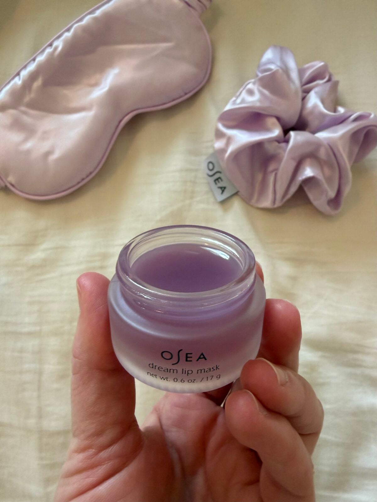 A hand holds an open jar of OSEA dream lip mask with light purple contents. In the background, there is a matching lavender sleep mask and a scrunchie placed on a cream-colored surface.