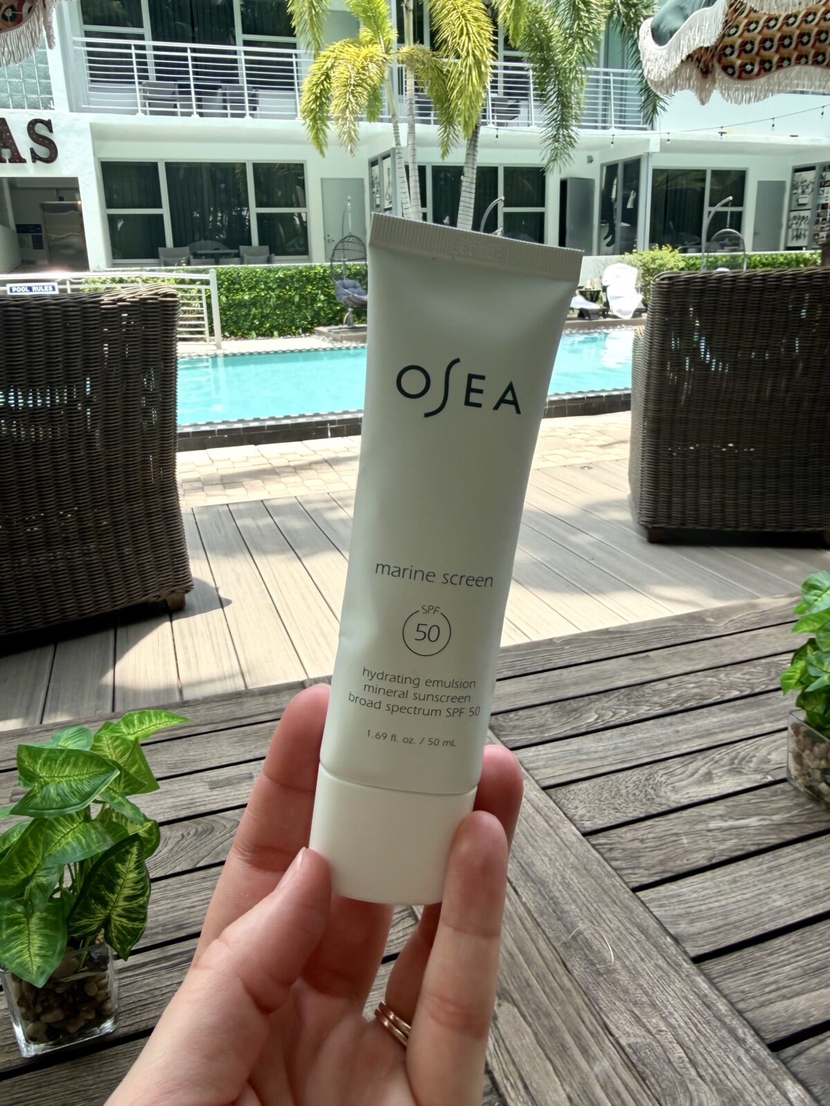A hand holds a tube of OSEA Marine Screen SPF 50 sunscreen outdoors by a pool, with wicker chairs, a wooden deck, potted plants, and a building with balconies in the background.