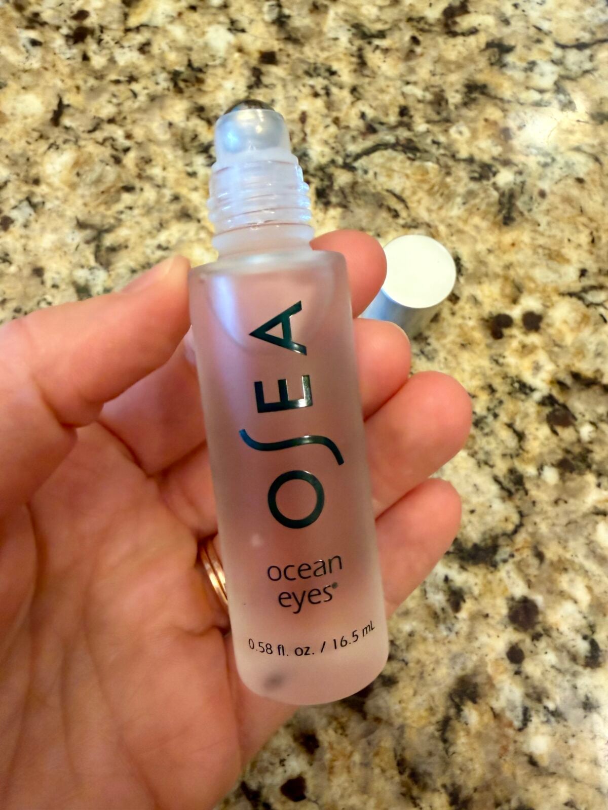 A hand holds a frosted glass bottle labeled OSEA ocean eyes with a rollerball applicator and a white cap, against a granite countertop background.