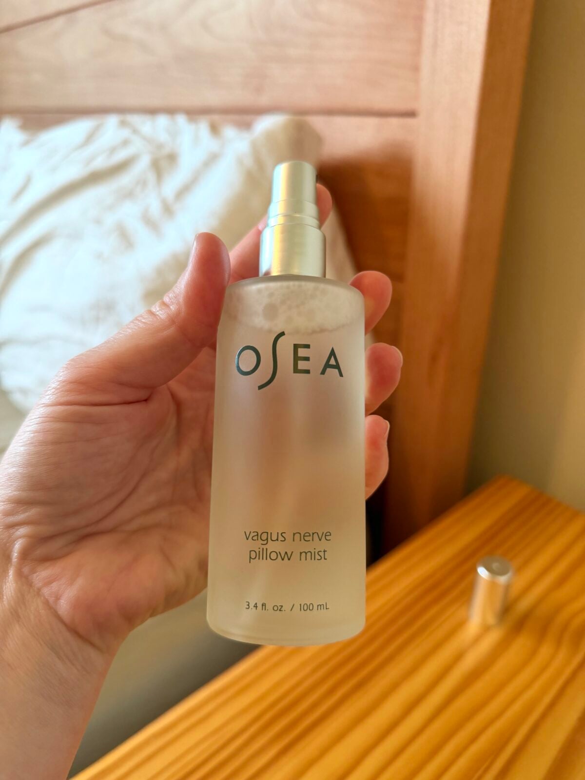 A hand holding a frosted glass spray bottle labeled OSEA vagus nerve pillow mist 3.4 fl. oz. / 100 mL next to a wooden nightstand, with a bed and bedding in the background.
