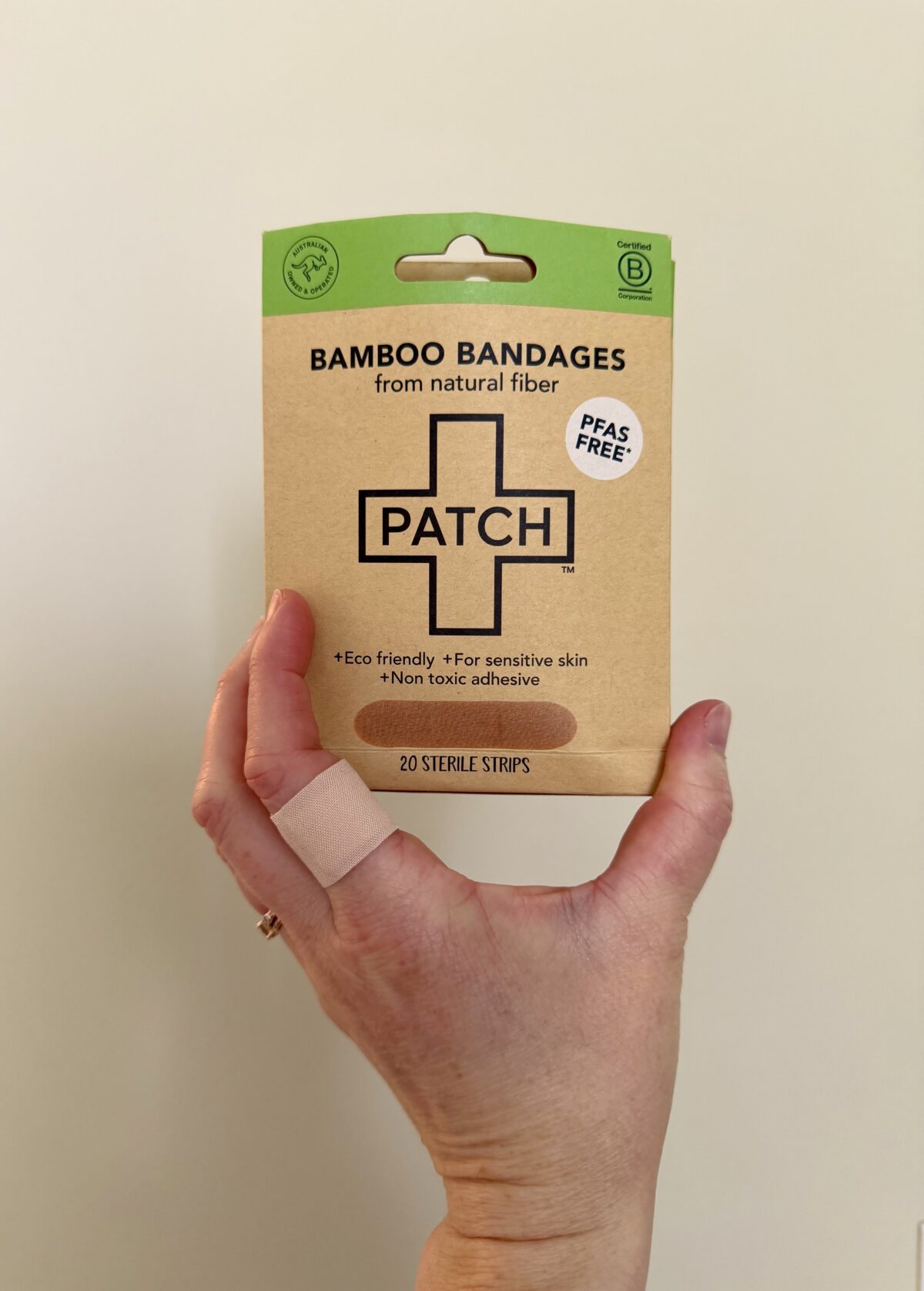A hand with a bandage on the index finger holds a box labeled “PATCH Bamboo Bandages from natural fiber.” The packaging states it is eco-friendly, suitable for sensitive skin, non-toxic, and contains 20 sterile strips.