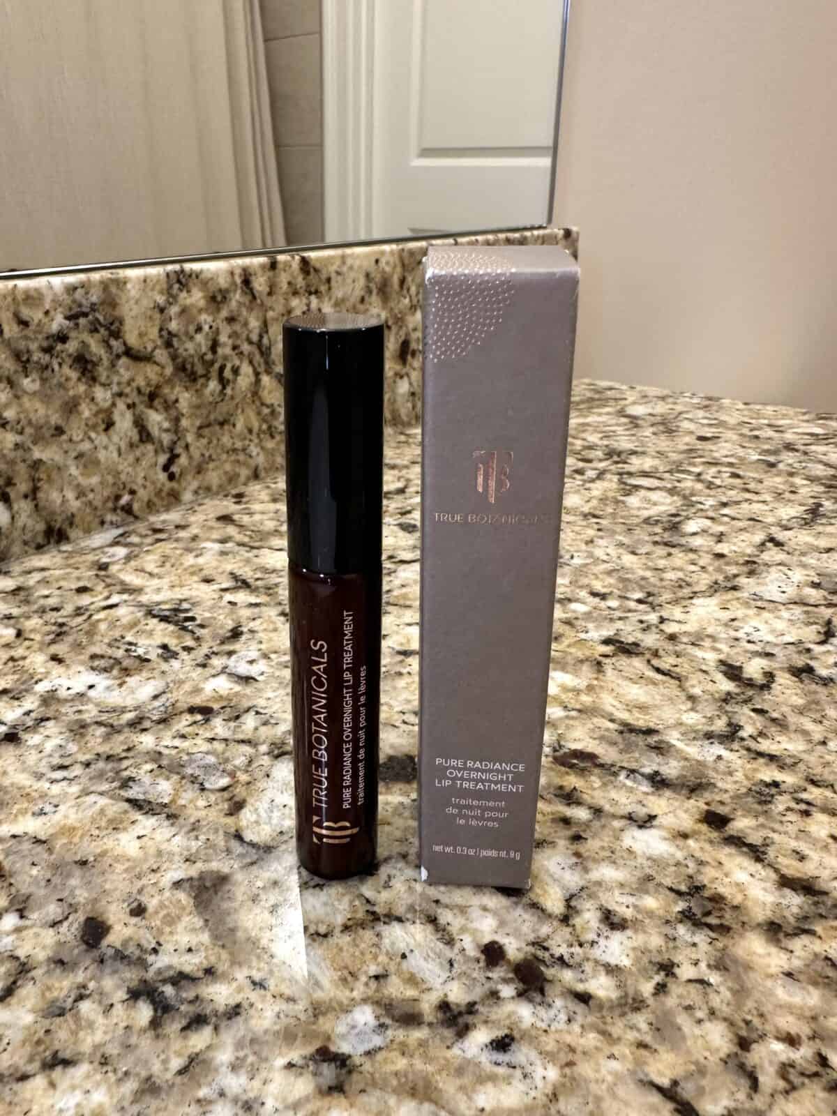 The photo shows the Pure Radiance Overnight Lip Treatmentfrom True Botanicals with its box arranged on a granite countertop with a bathroom mirror in the background.