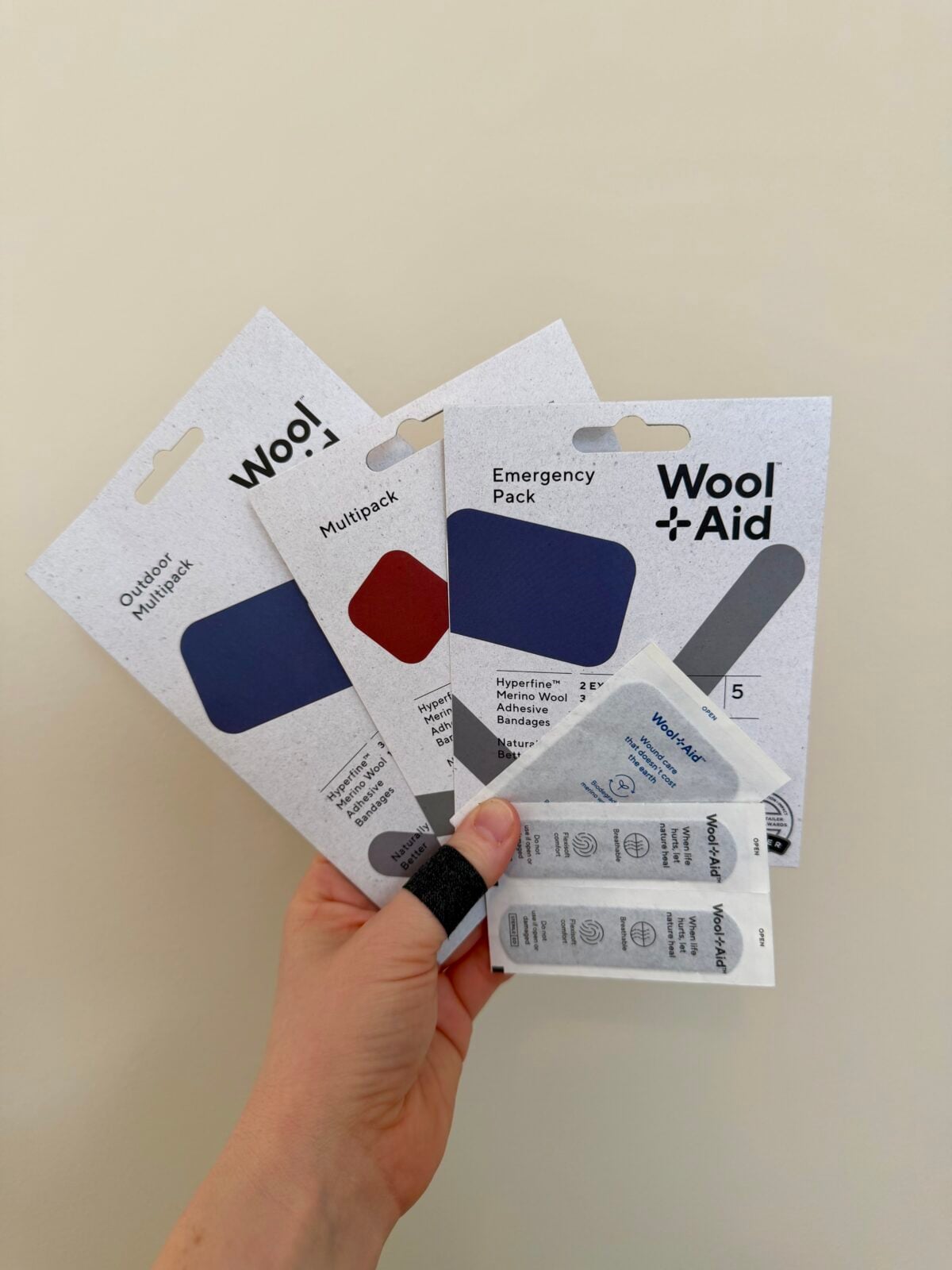 A hand holds three WoolAid bandage packs and two single-use antiseptic wipe packets against a light beige background. One finger on the hand is wrapped with a dark bandage.