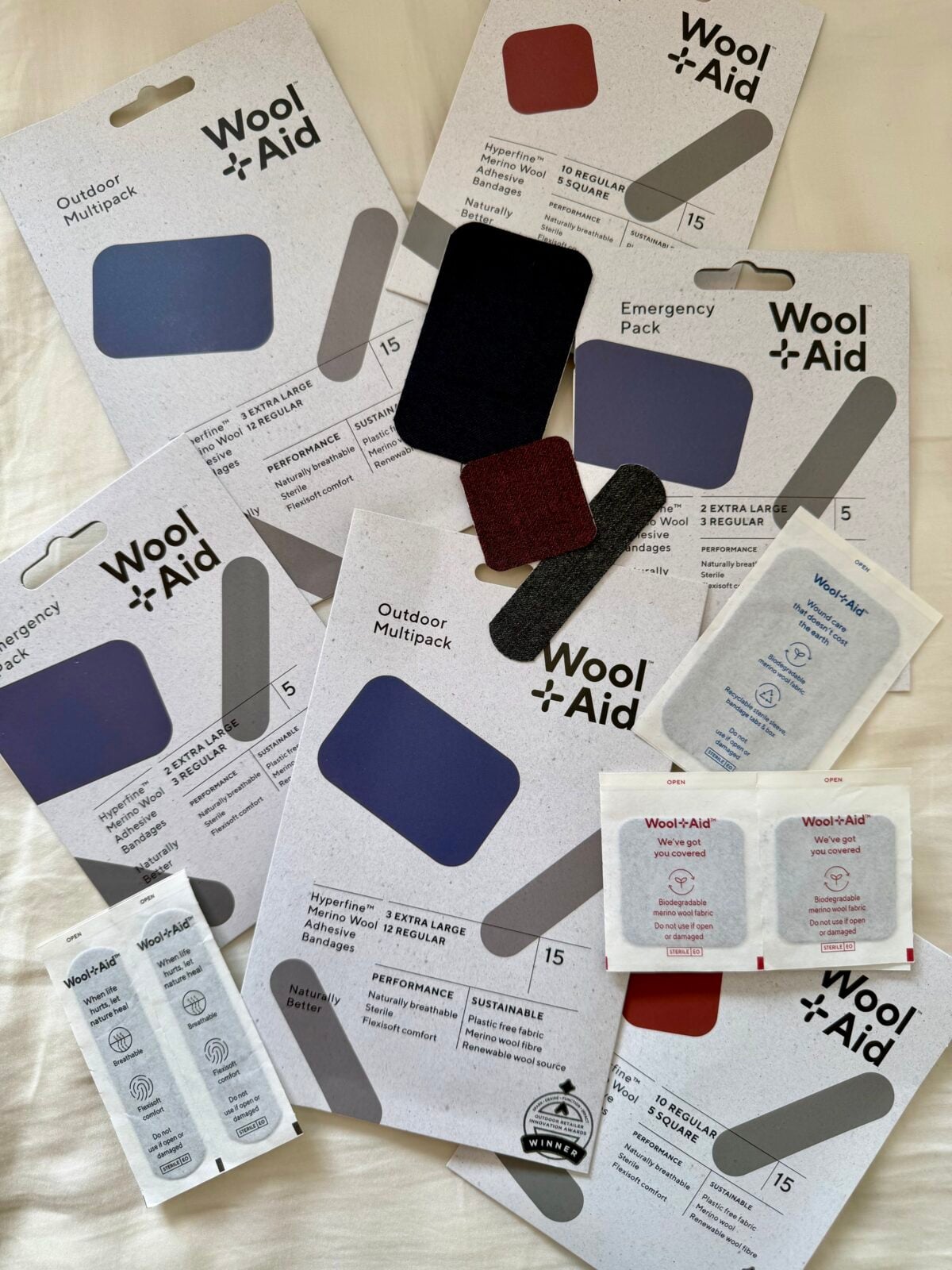 Several Wool+Aid branded wound care products are spread out on a white surface, including Outdoor and Emergency packs, colorful adhesive patches, antiseptic wipes, and single-use cream packets.
