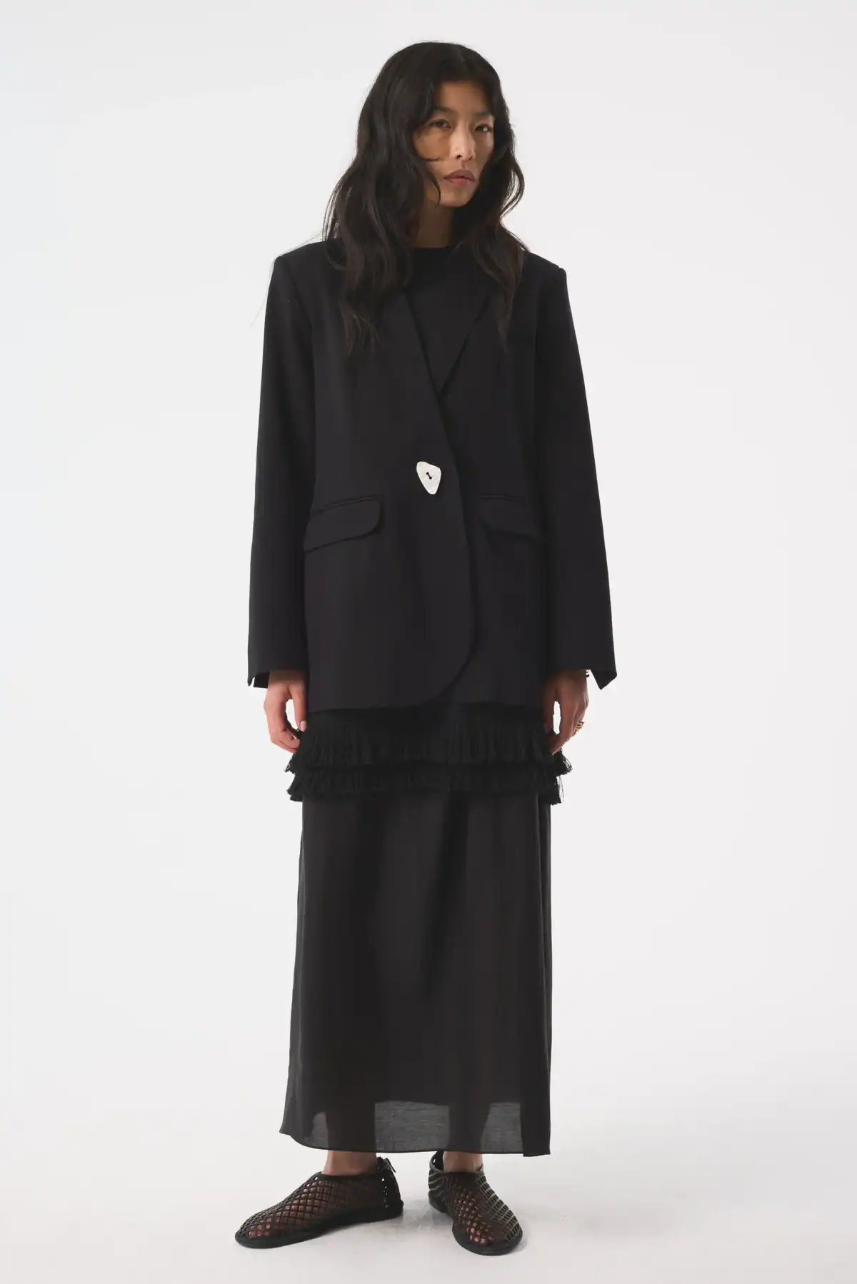 A person with long dark hair is standing against a white background, wearing a black blazer from Akyn, over a sheer black ruffle skirt and black shoes.
