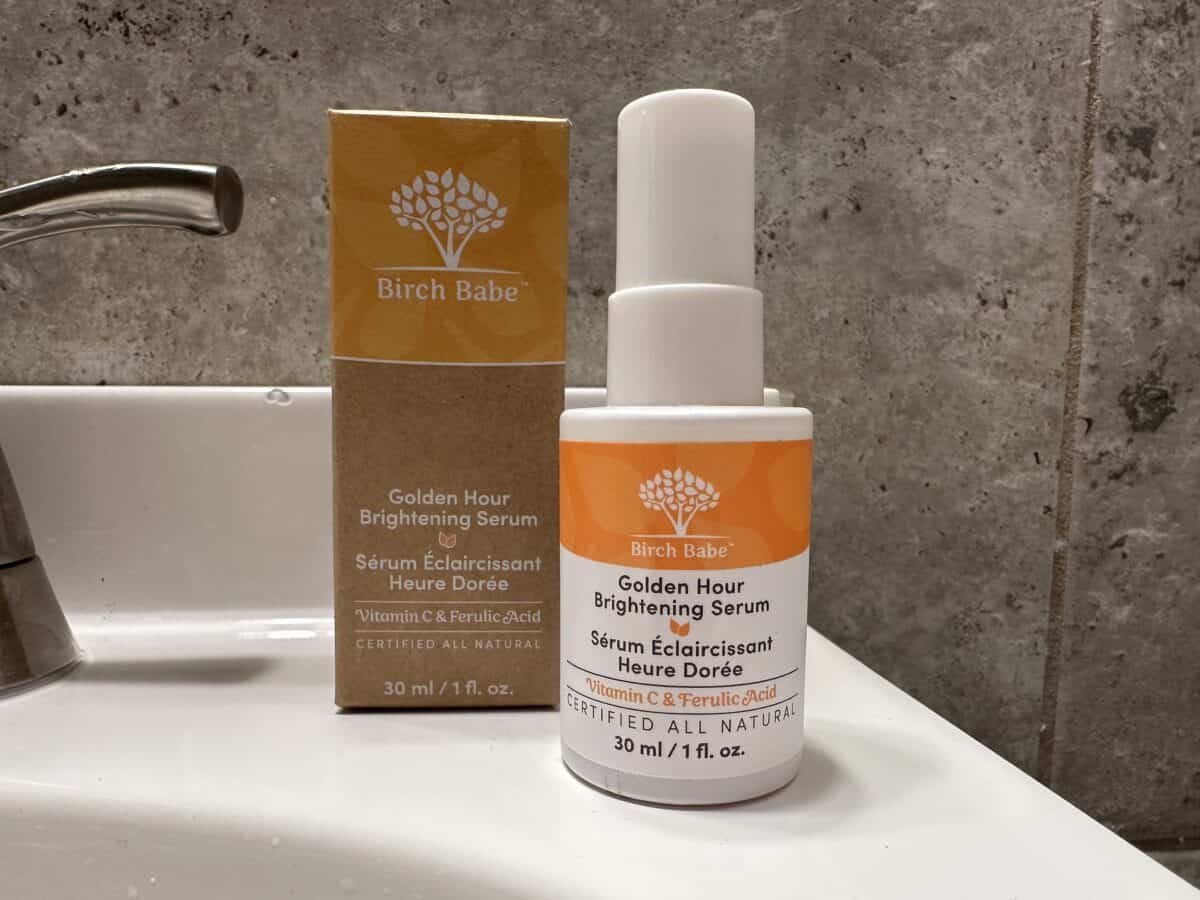 The photo features a bottle and box of Birch Babe "Golden Hour Brightening Serum" positioned on a white bathroom sink next to a silver faucet.